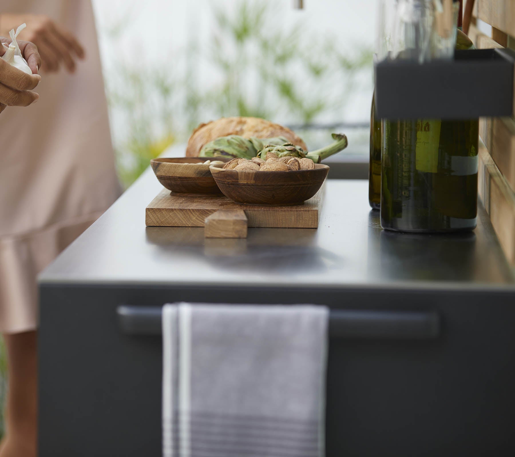 Eine Nahaufnahme einer Küchenarbeitsplatte mit Holtschalen voller Essen, einem Handtuch und Flaschen mit Olivenöl, mit sanften Pflanzen im Hintergrund.