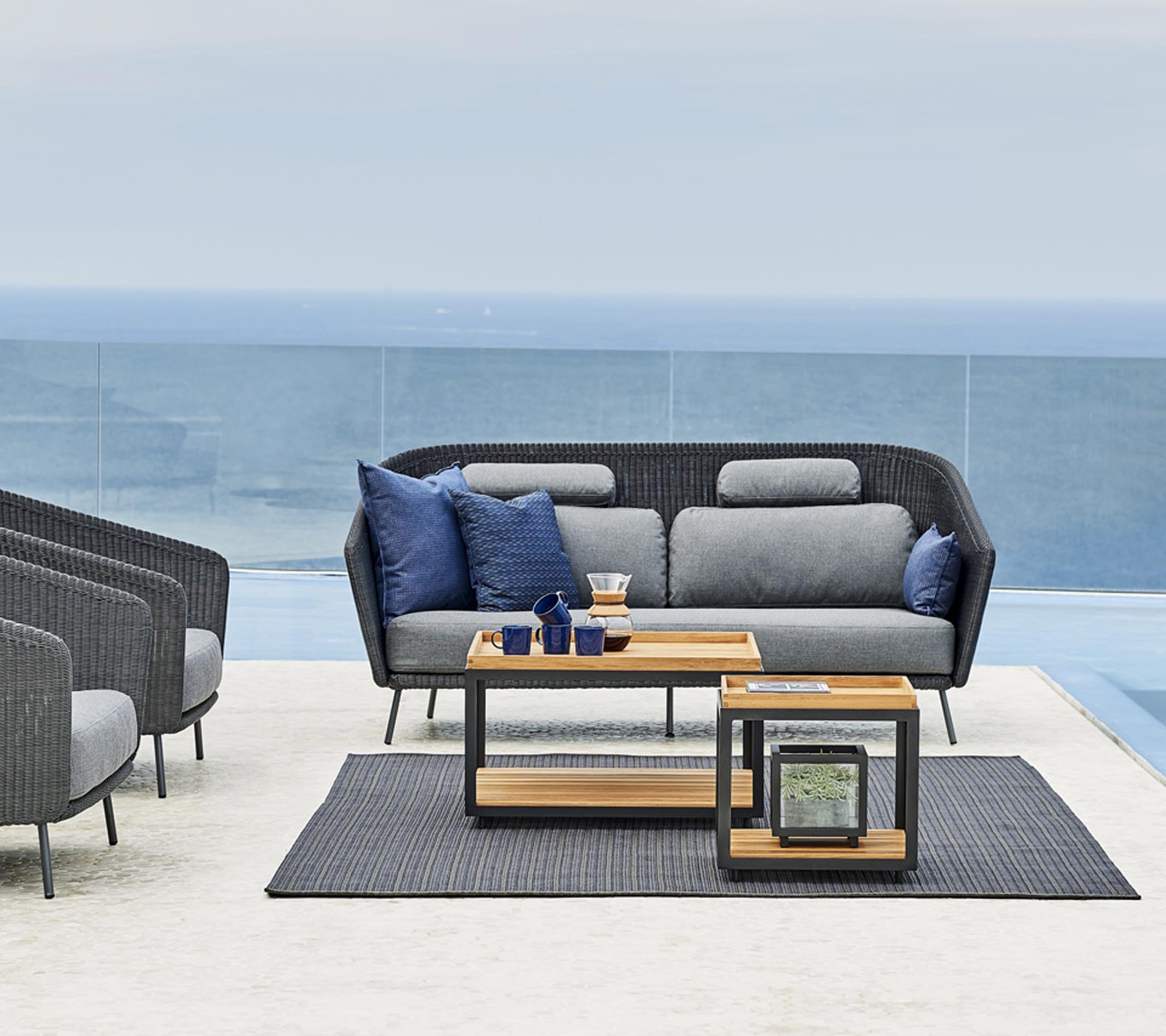 Stylische Außensitzanordnung mit einem grauen Sofa mit blauen Kissen, einem Holztisch und einem passenden Teppich.