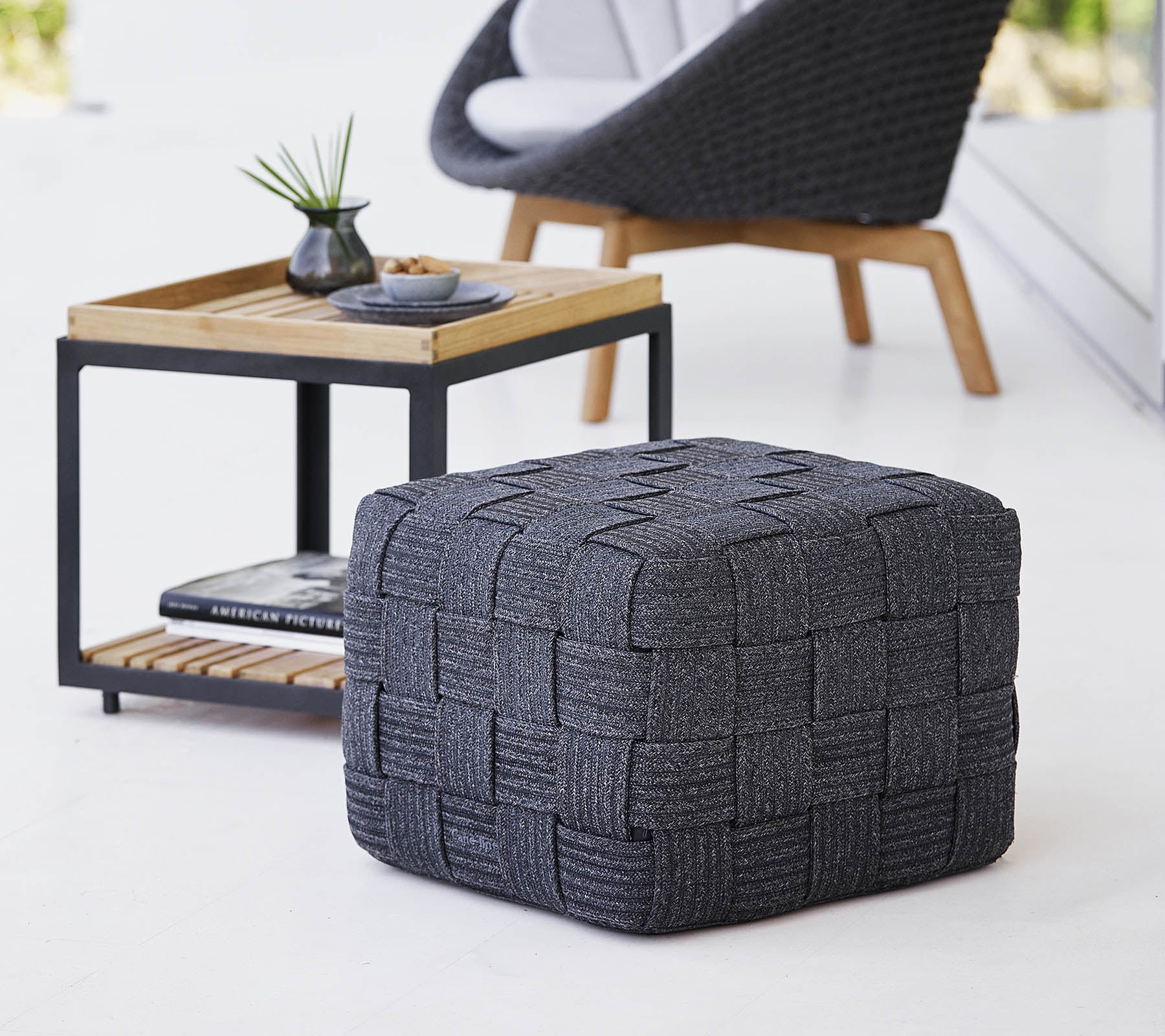 Ein strukturierter grauer gewebter Pouf, der neben einem minimalistischen Holztisch steht, der kleine Dekorationsartikel enthält.