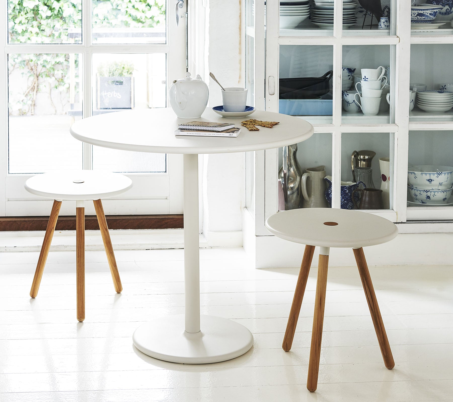 Ein runder weißer Tisch, begleitet von zwei minimalistischen Hockern mit Holzbeinen, vor einem Hintergrund aus Regalen mit Küchenutensilien.