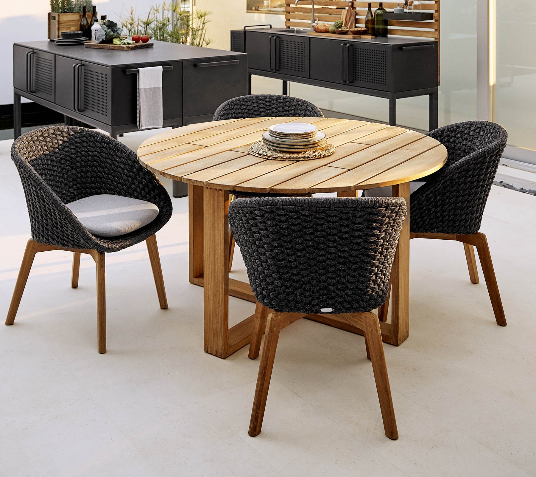 Runder Holztisch mit vier stilvollen geflochtenen Stühlen, perfekt für Dinner und gesellschaftliche Zusammenkünfte. Elegante und einladende Sitzgelegenheit im Freien.