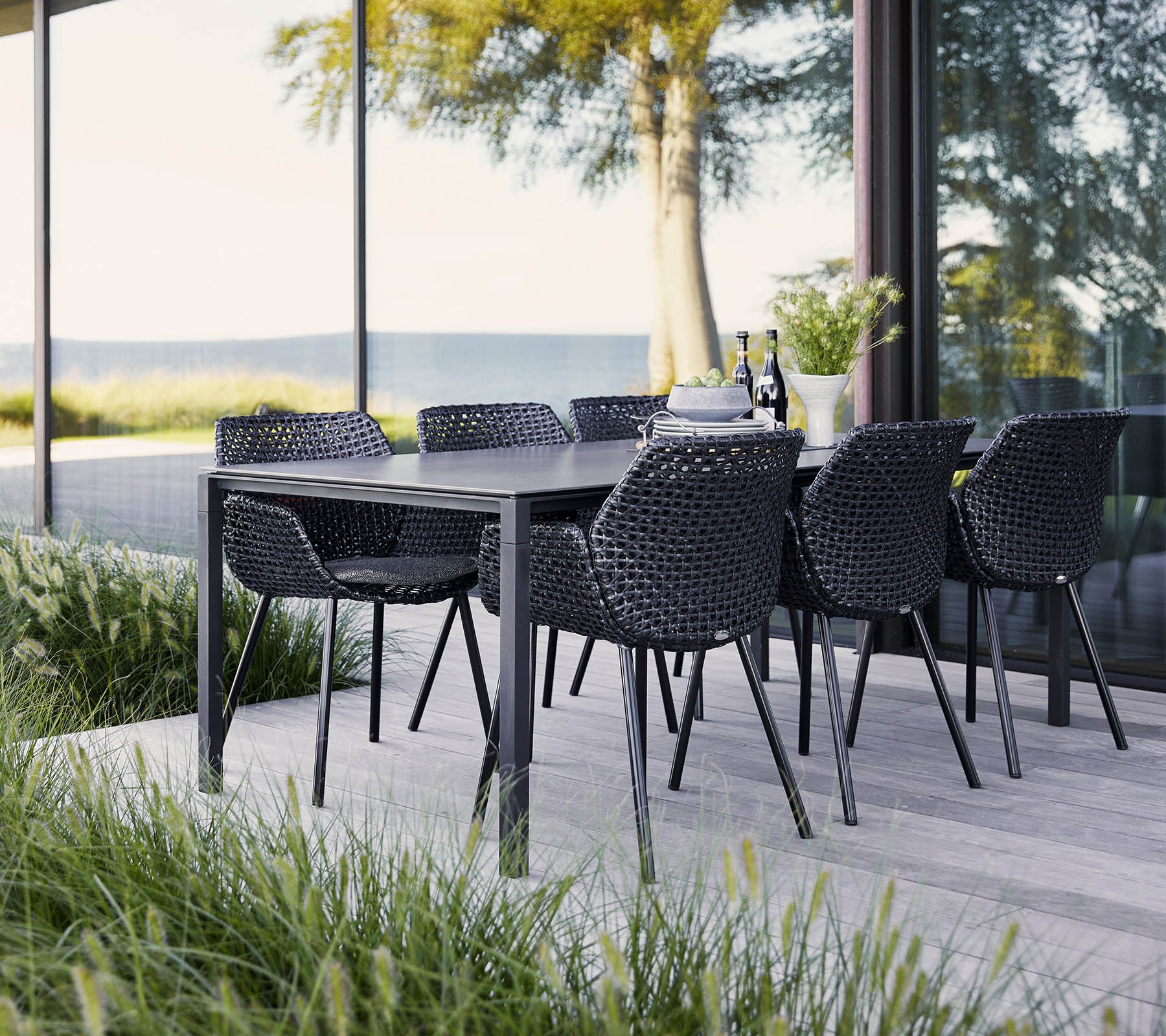 Ein stilvolles Essset mit einem schwarzen Tisch und geflochtenen Stühlen, umgeben von üppigem Grün und weitem Ausblick. Ideal für Zusammenkünfte.