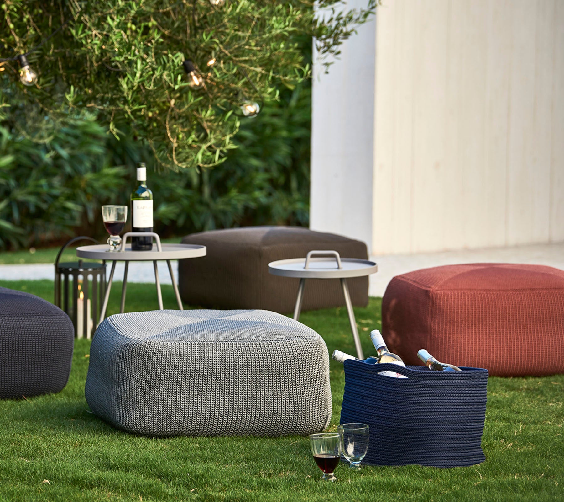 Texturierte Poufs in verschiedenen Farben auf dem Gras angeordnet, begleitet von einem kleinen Tisch mit Getränken und Weinflaschen.