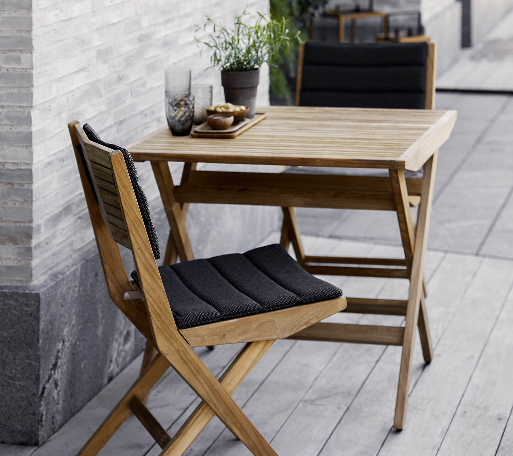 Holztisch und Stuhlset mit schwarzen Kissen, geschmückt mit Topfpflanzen und dekorativen Gläsern. Perfekt für eine gemütliche Atmosphäre.