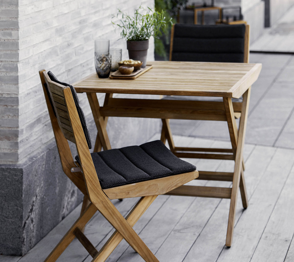 Holztisch und Stuhlset mit schwarzen Kissen, geschmückt mit Topfpflanzen und dekorativen Gläsern. Perfekt für eine gemütliche Atmosphäre.