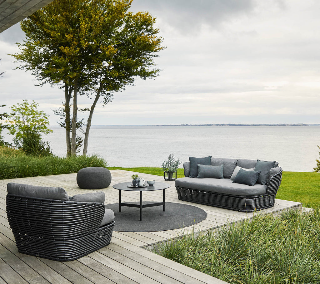 Gemütliche Sitzgelegenheit mit einem Sofa und Stühlen, die um einen runden Tisch angeordnet sind, umgeben von Grün und mit Blick auf das Wasser.