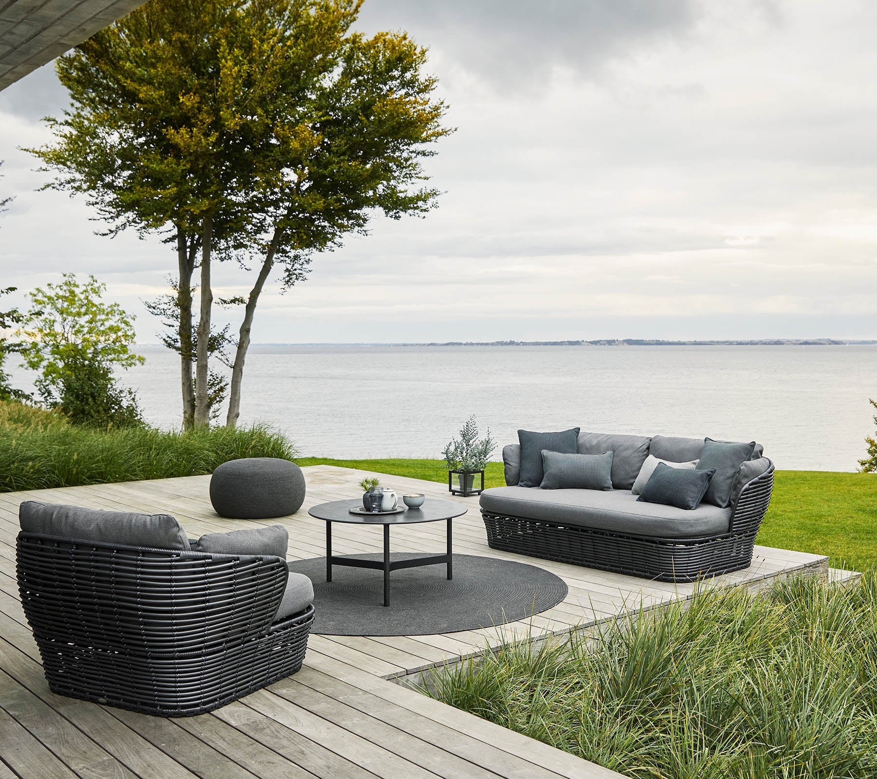 Gemütliche Sitzgelegenheit mit einem Sofa und Stühlen, die um einen runden Tisch angeordnet sind, umgeben von Grün und mit Blick auf das Wasser.