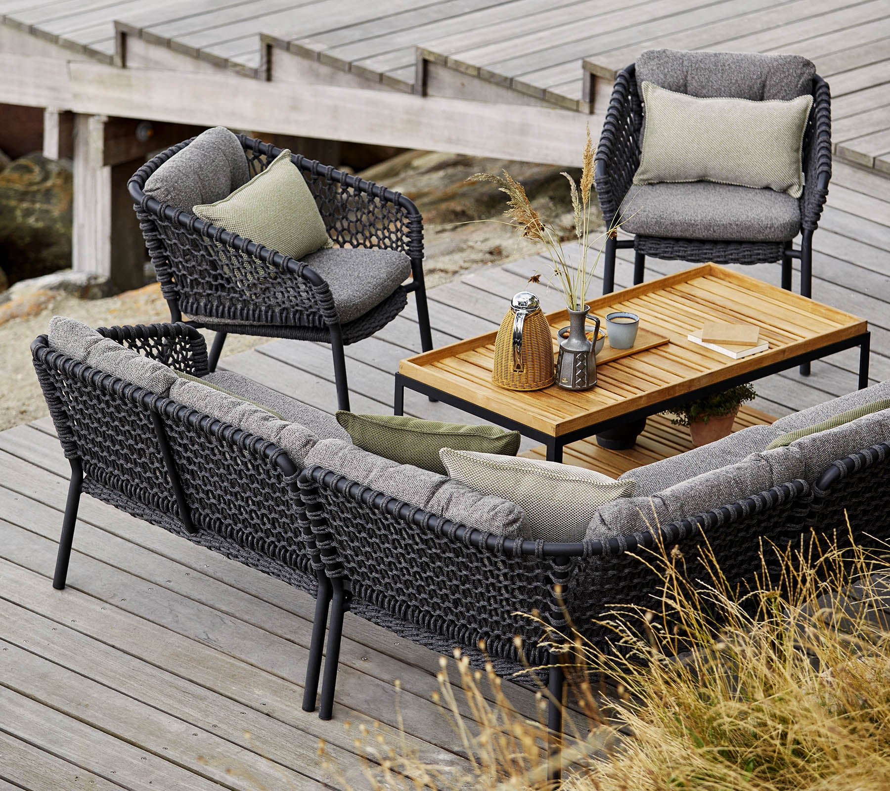 Stilvolles Outdoor-Sitzset mit einer gemütlichen Couch, zwei Stühlen und einem Holztisch, akzentuiert mit weichen Kissen und Dekorationsgegenständen.