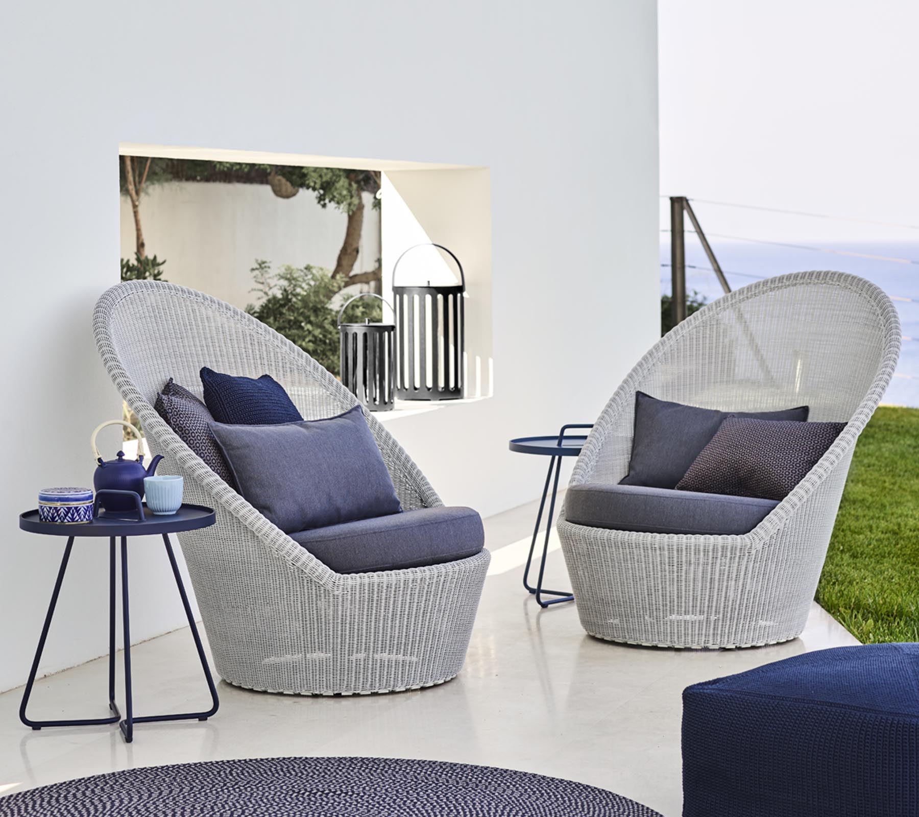 Zwei stilvolle, geflochtene Lounge-Stühle mit blauen Kissen neben einem kleinen Tisch, vor einer ruhigen Kulisse.