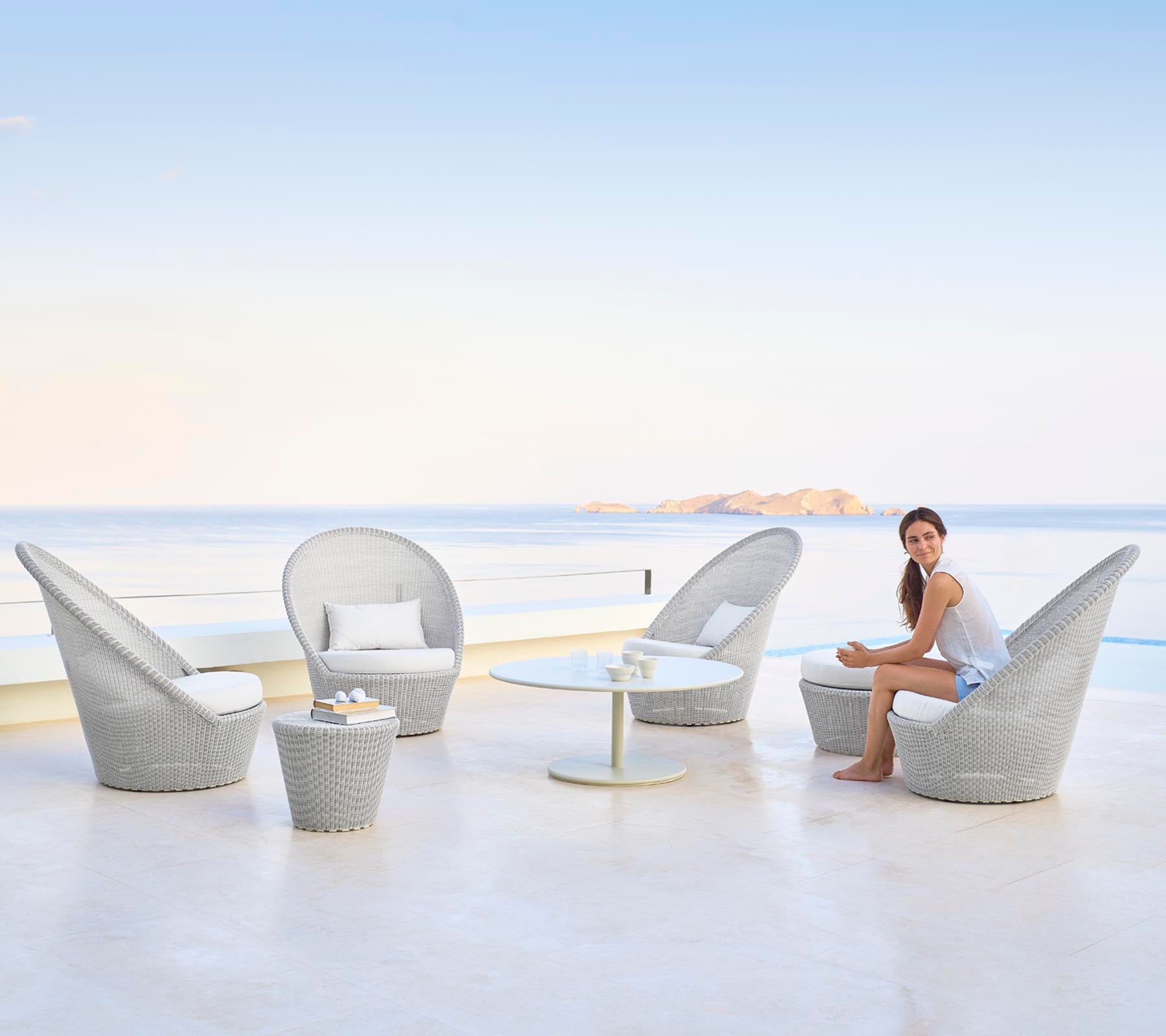 Eine stilvolle Sitzanordnung mit eleganten Stühlen und einem Tisch, an dem eine Frau anmutig vor einer ruhigen Wasserlandschaft sitzt.
