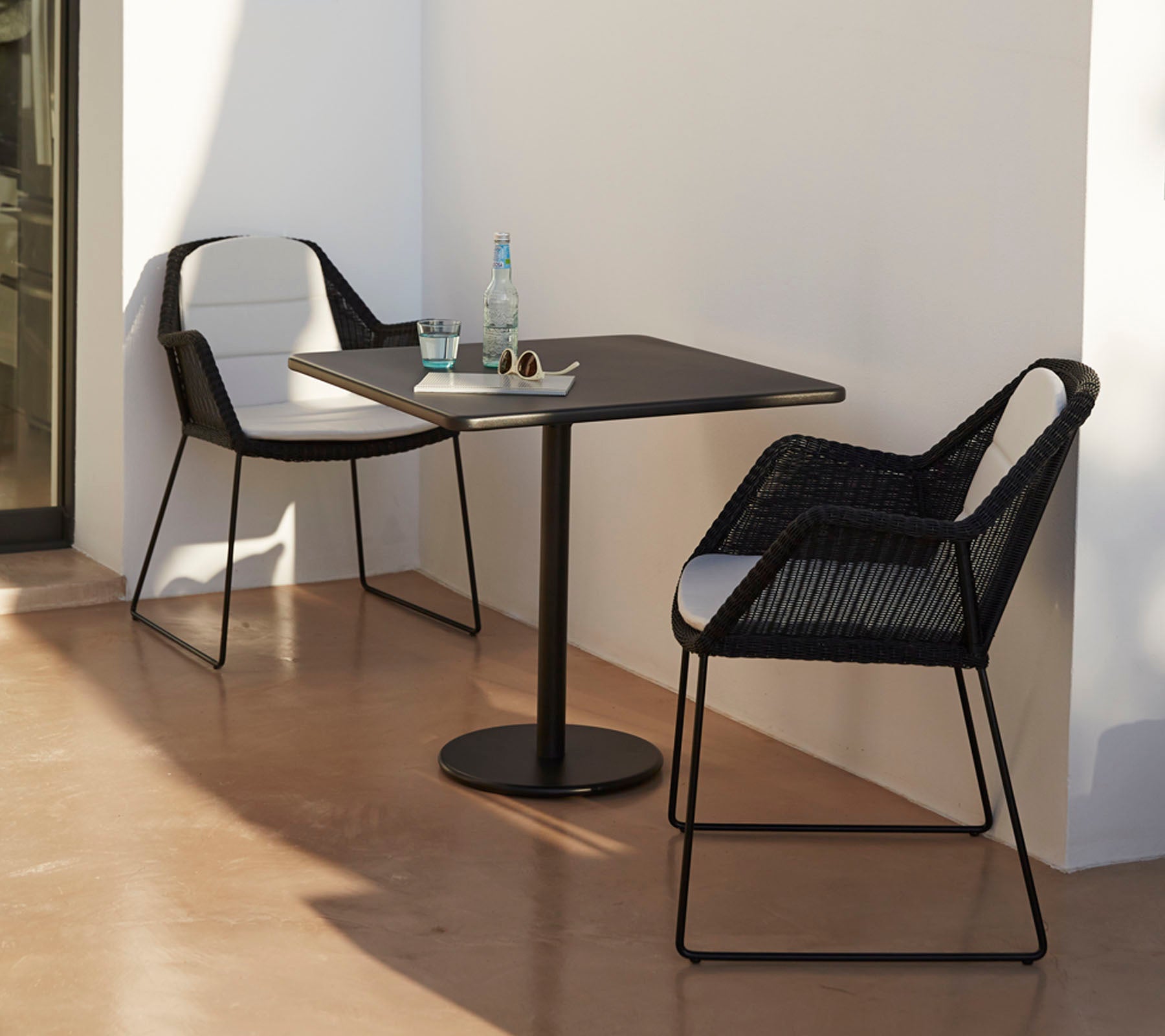 Modernes Tisch mit zwei stilvollen schwarzen Stühlen, begleitet von einer Wasserflasche und Sonnenbrille, vor einem minimalistischen Hintergrund.