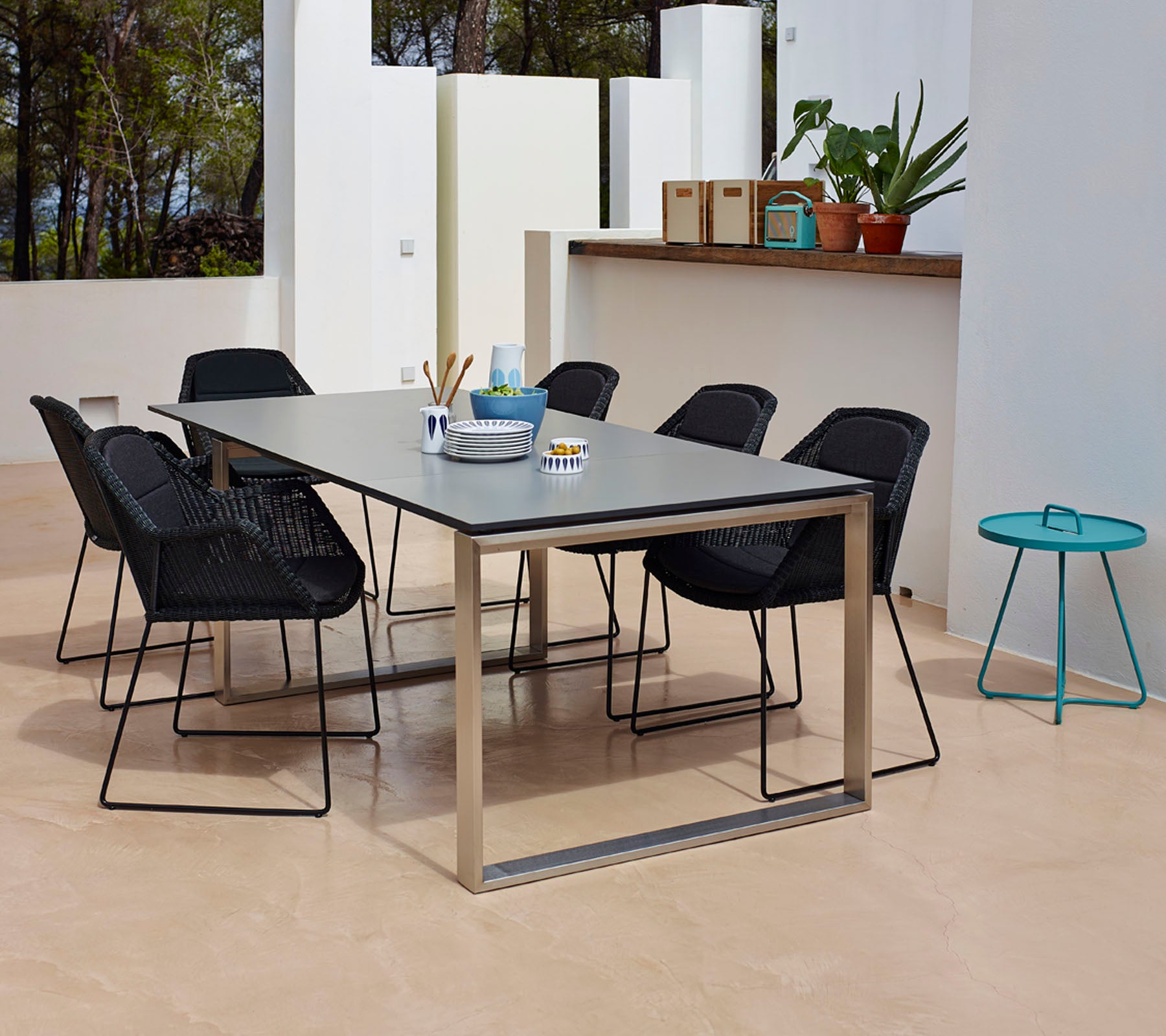 Ein modernes Esszimmer-Setup mit einem rechteckigen Tisch mit einer glatten Oberfläche, umgeben von sechs schwarzen geflochtenen Stühlen und einem kleinen türkisfarbenen Beistelltisch.