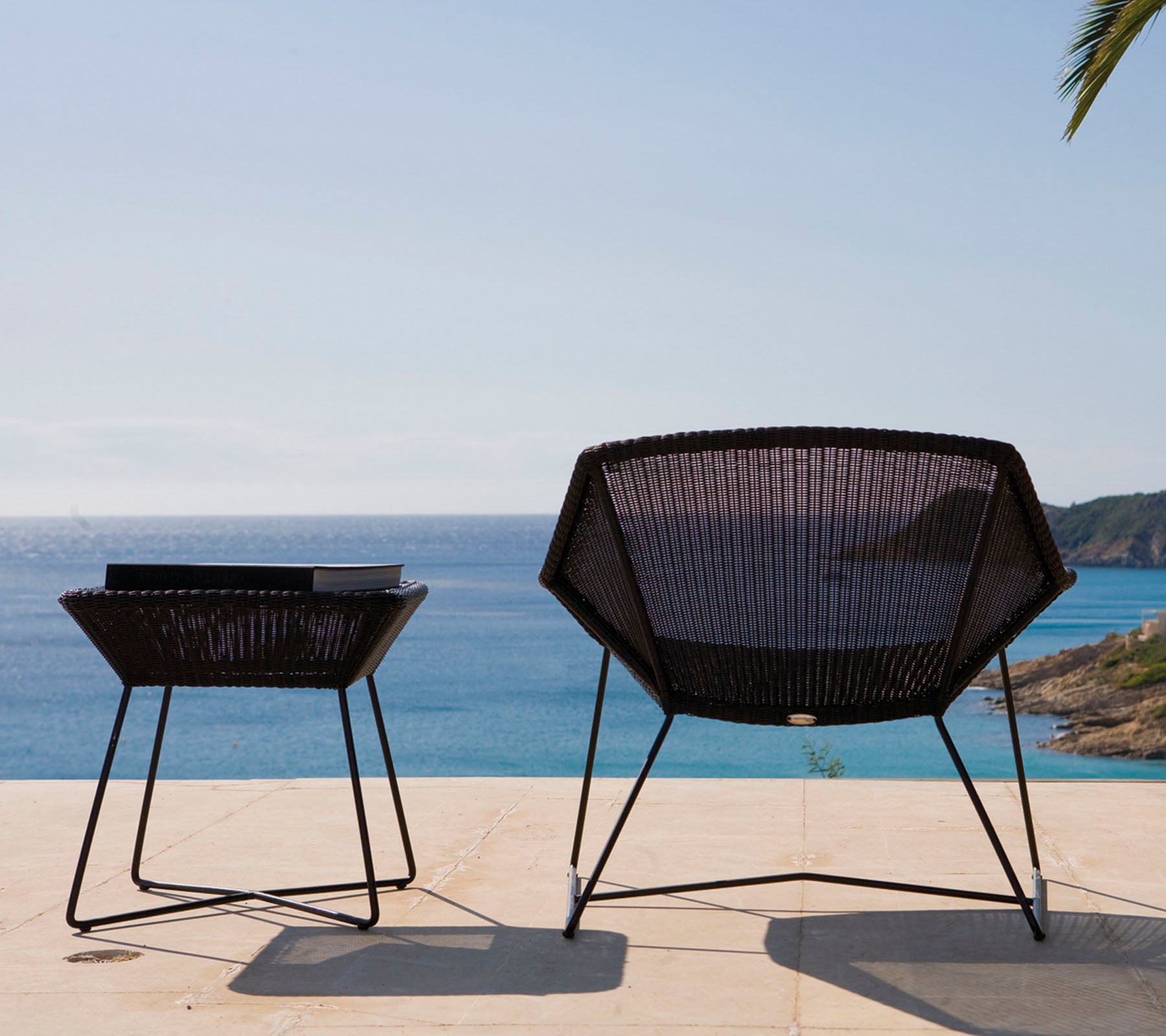 Zwei moderne, schwarze Gartensessel mit einem geometrischen Design stehen neben einem Tisch und blicken auf ein ruhiges Meer unter einem klaren Himmel.