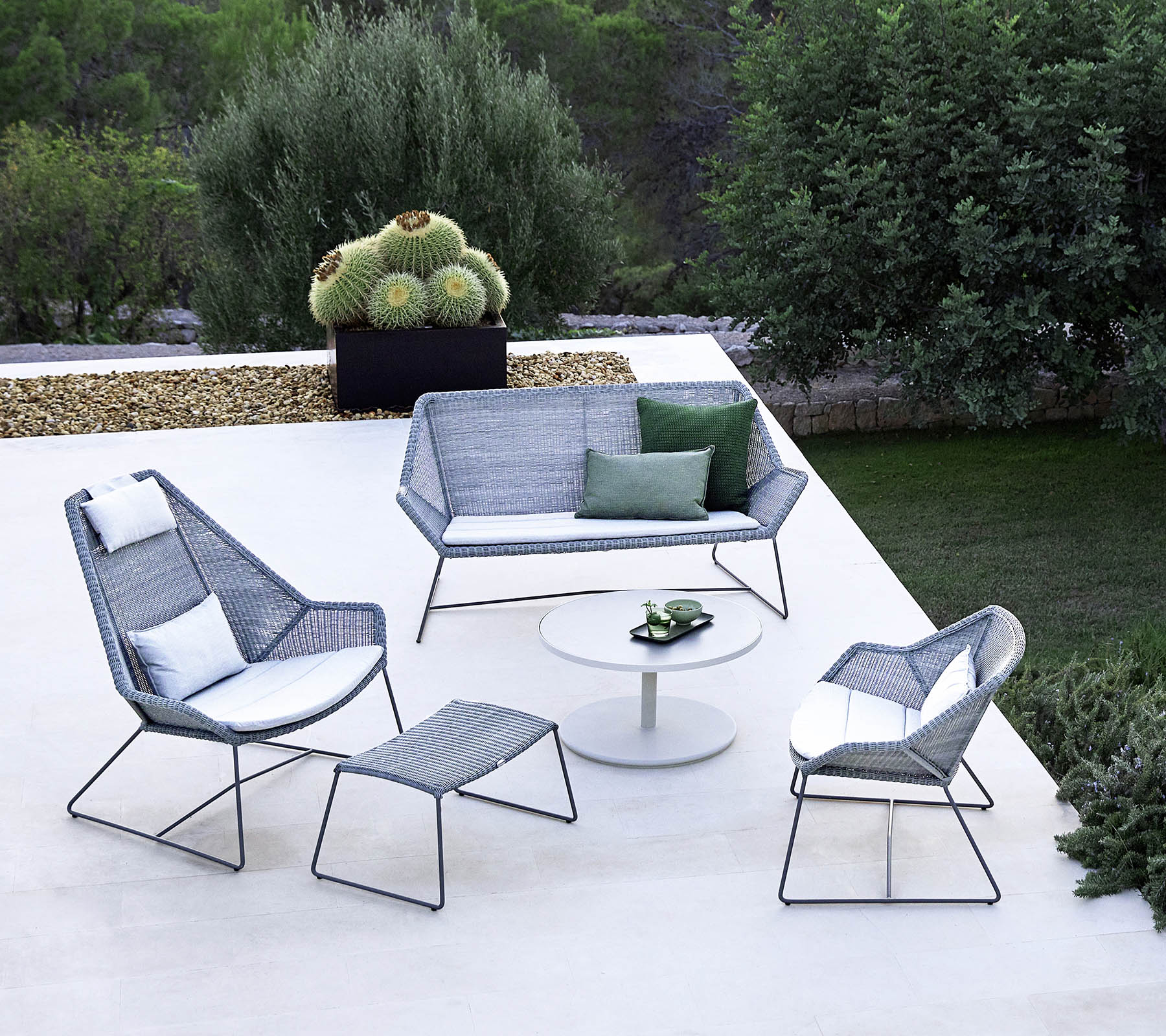 Stylisches Außen-Sitzarrangement mit einem Sofa, zwei Armlehnstühlen, einem Fußhocker und einem Couchtisch, ergänzt durch Grünpflanzen im Hintergrund.