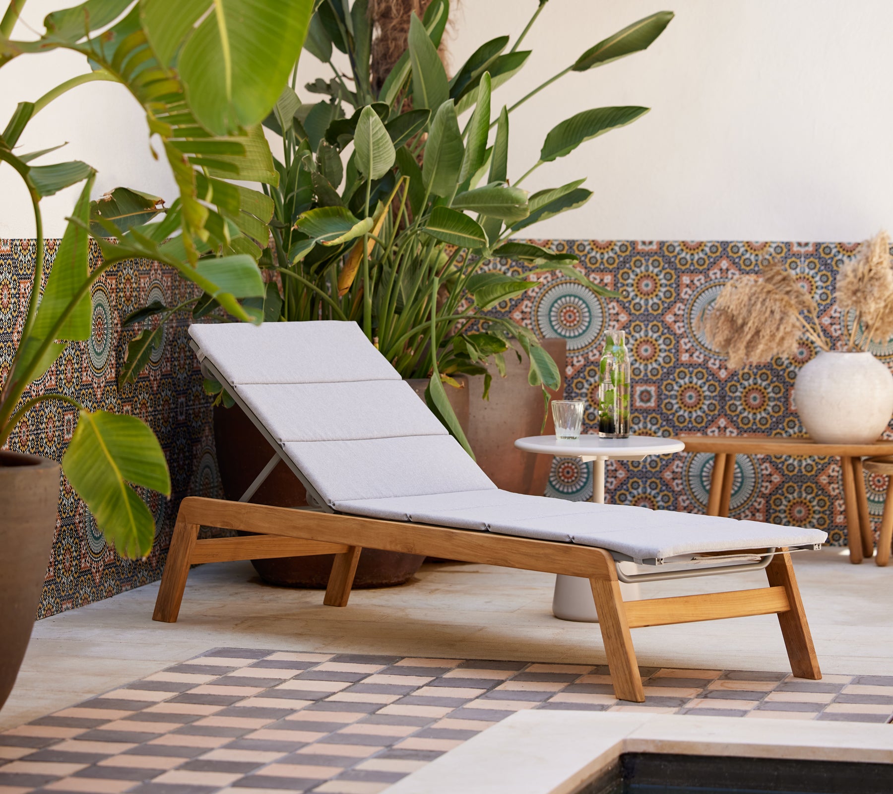Ein moderner Lounge-Stuhl mit einem leichten Kissen, kombiniert mit einem Beistelltisch, umgeben von üppigem Grün und dekorativen Fliesen im Hintergrund.
