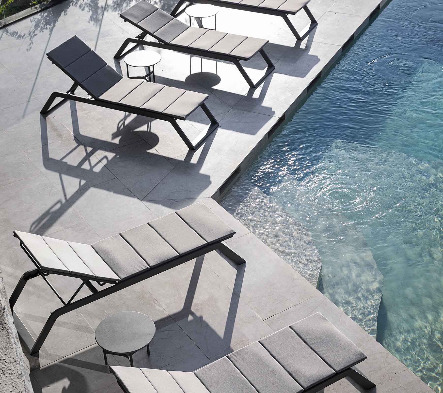 Eine Reihe moderner Lounge-Stühle mit einem kleinen Beistelltisch neben einem Swimmingpool, der Schatten auf das Pflaster wirft.