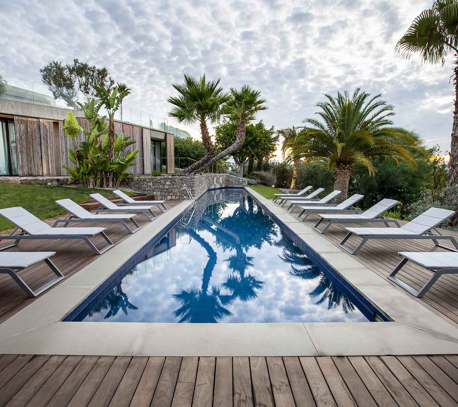 Serene Szene mit einem Pool, umgeben von Liegen und Palmen, die den Himmel und das üppige Grün reflektiert.