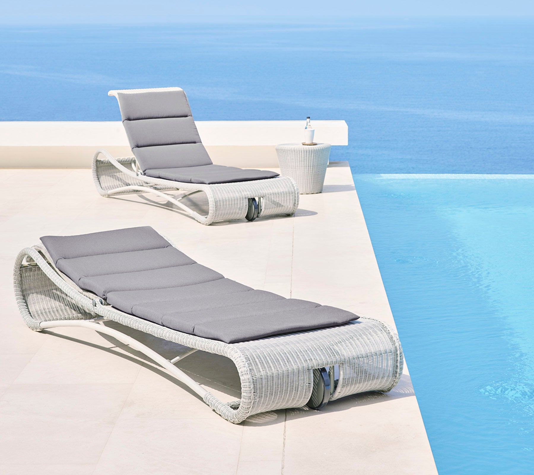 Zwei stilvolle Loungestühle mit grauen Kissen neben einem ruhigen blauen Infinity-Pool, die einen entspannenden Außenbereich schaffen.