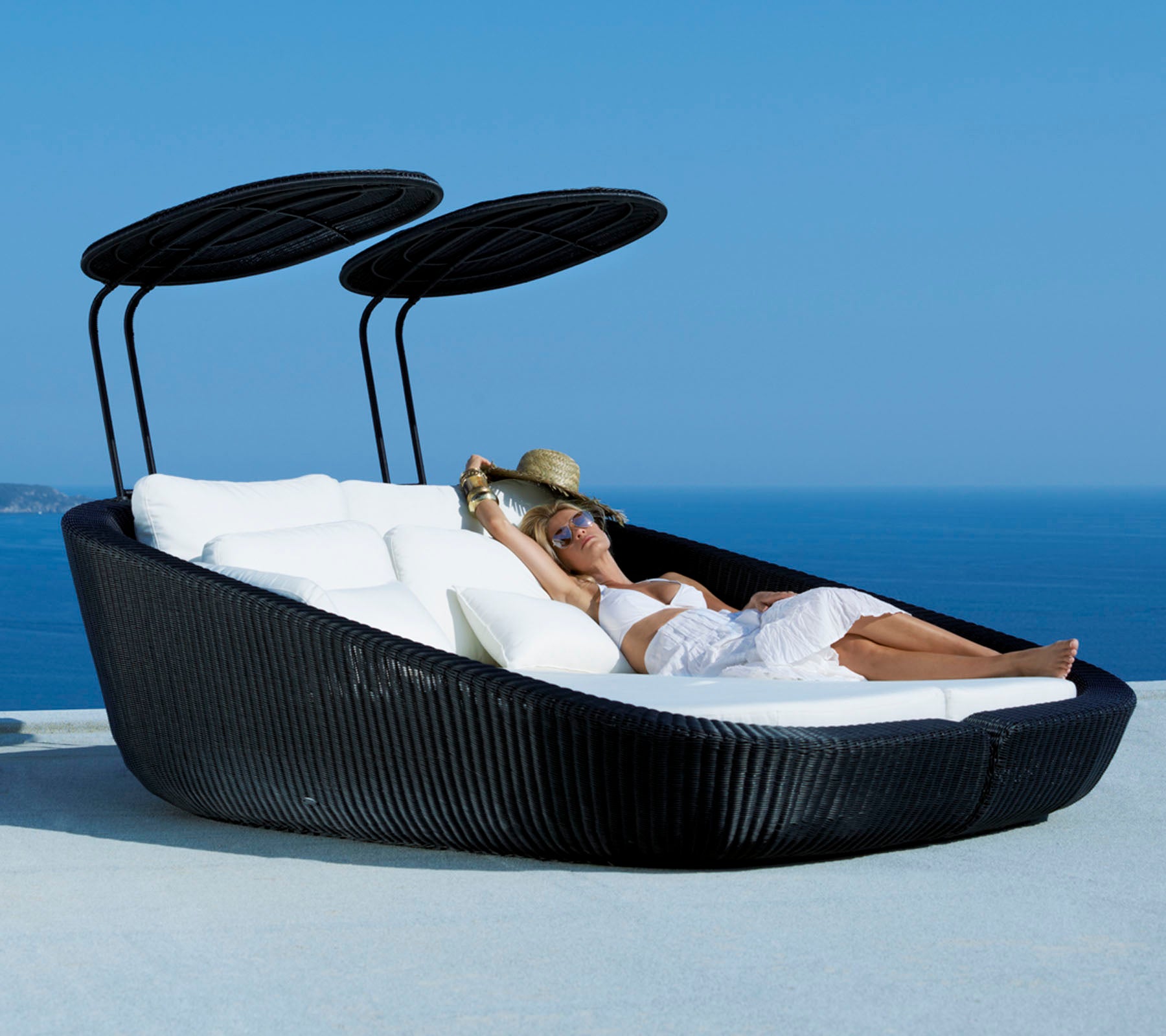 Ein stilvolles Loungesofa für den Außenbereich mit einem schlanken Design, das weiche Kissen und verstellbare Sonnendächer bietet, vor einer malerischen Ozeankulisse.
