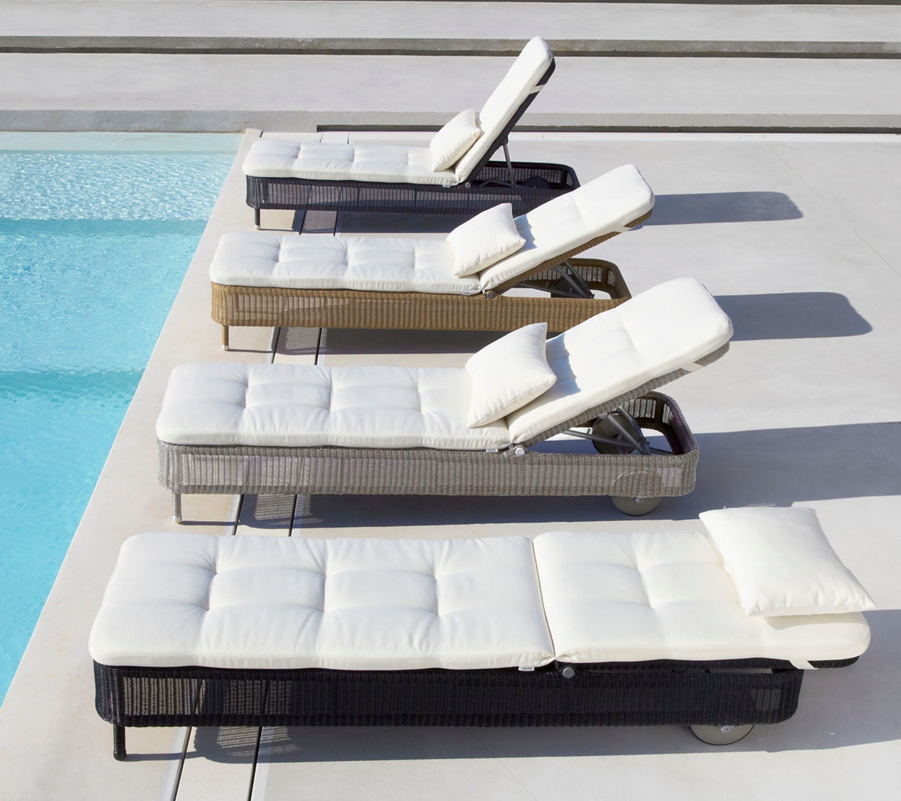 Vier Liegen in verschiedenen Farben mit weißen Kissen, neben einem Pool positioniert, jede mit verstellbarer Rückenlehne.