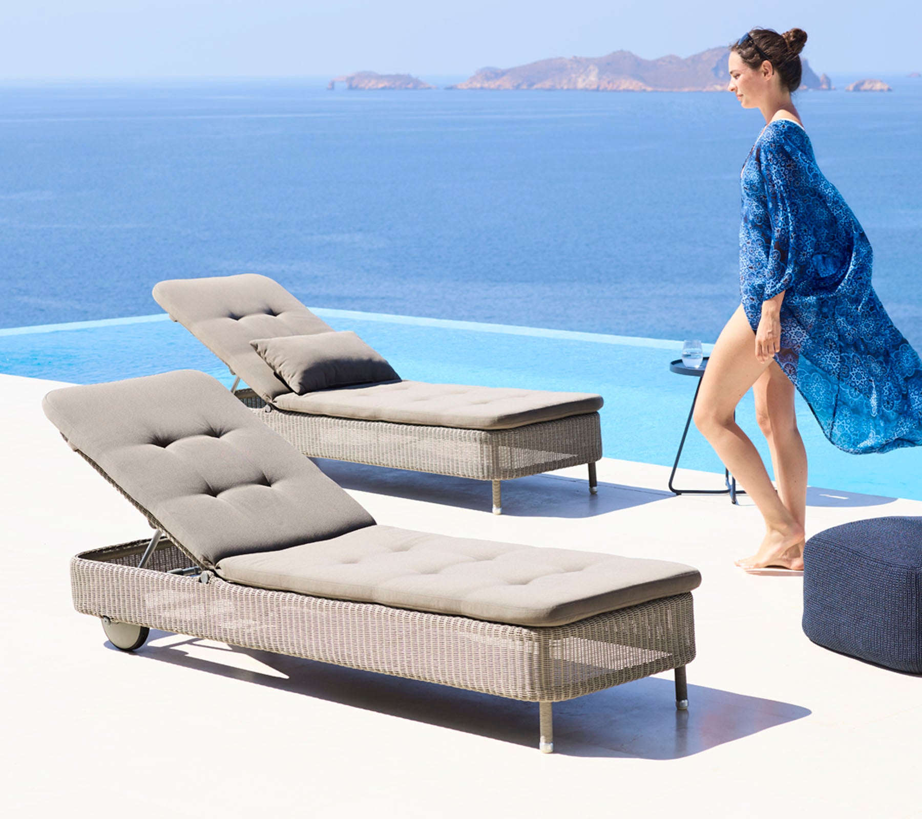 Eine Frau in einem fließenden blauen Cover-up geht neben zwei stilvollen Liegestühlen am Pool mit Blick auf einen ruhigen Ozean.