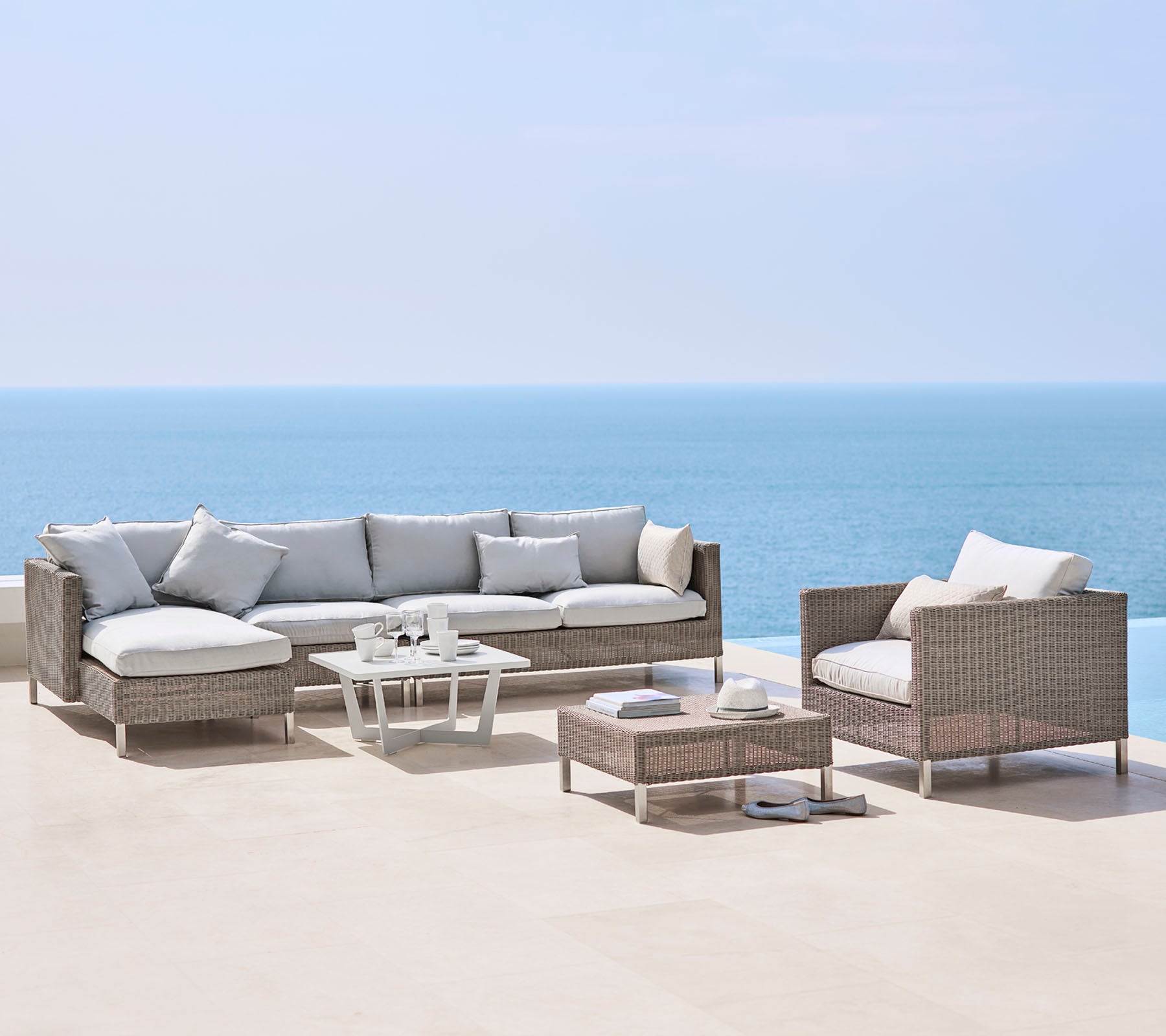 Eine stilvolle Outdoor-Sitzgarnitur mit einem großen Sofa, einem Sessel und einem Couchtisch, alle mit hellen Kissen und geflochtenem Material, am Meer.