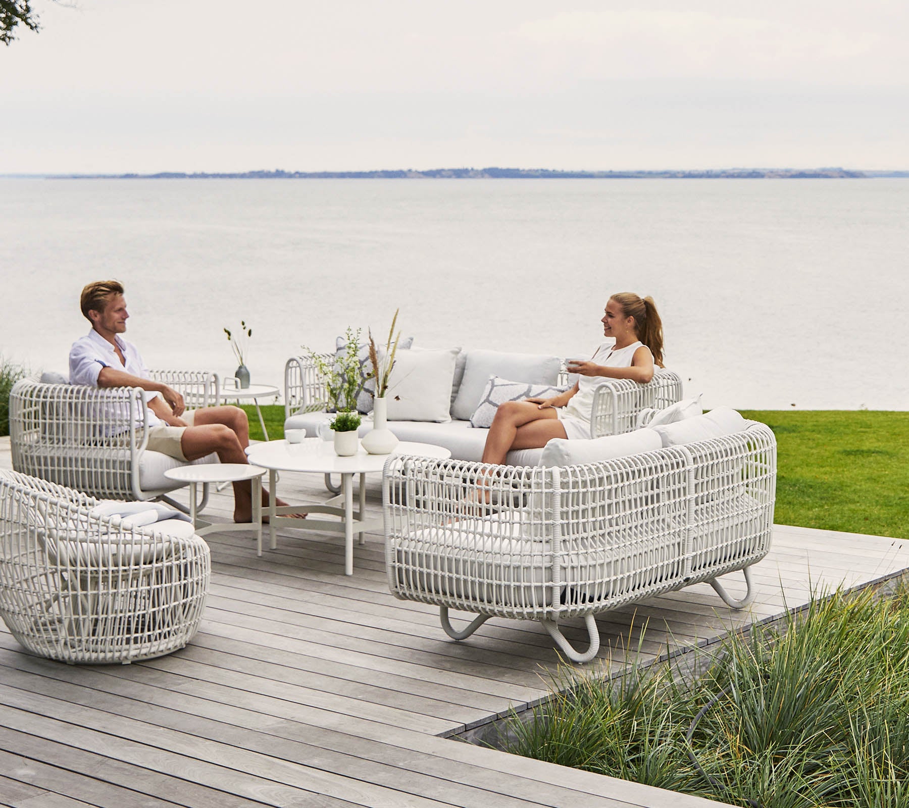 Zwei Personen entspannen sich in einem stilvollen, bequemen Sitzbereich mit weißen Korbmöbeln und einem Couchtisch, mit Blick auf eine malerische Wasserlandschaft.