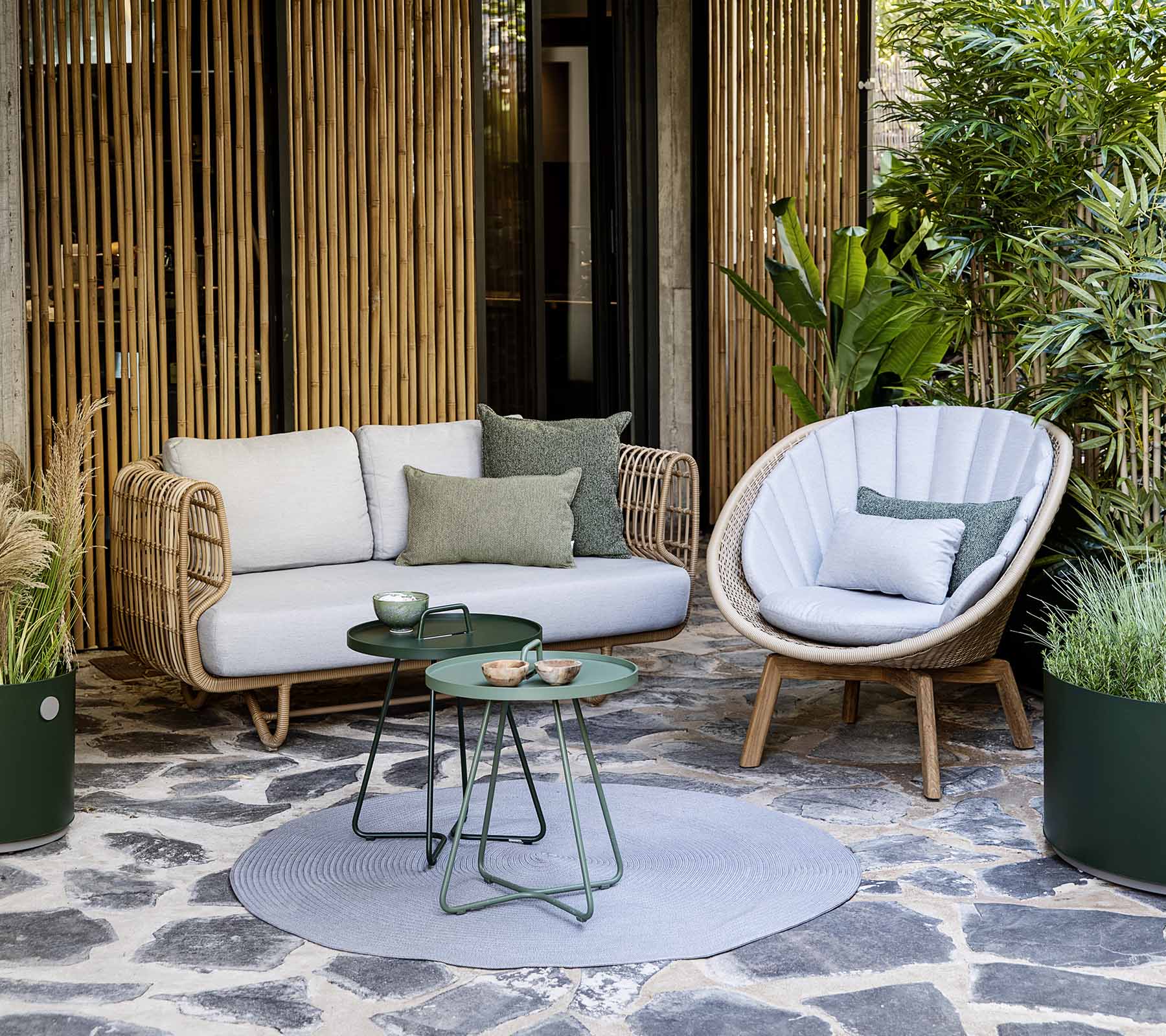 Eine gemütliche Sitzgelegenheit mit einem Rattansofa und einem stilvollen Stuhl, begleitet von einem kleinen Tisch und dekorativen Pflanzen.