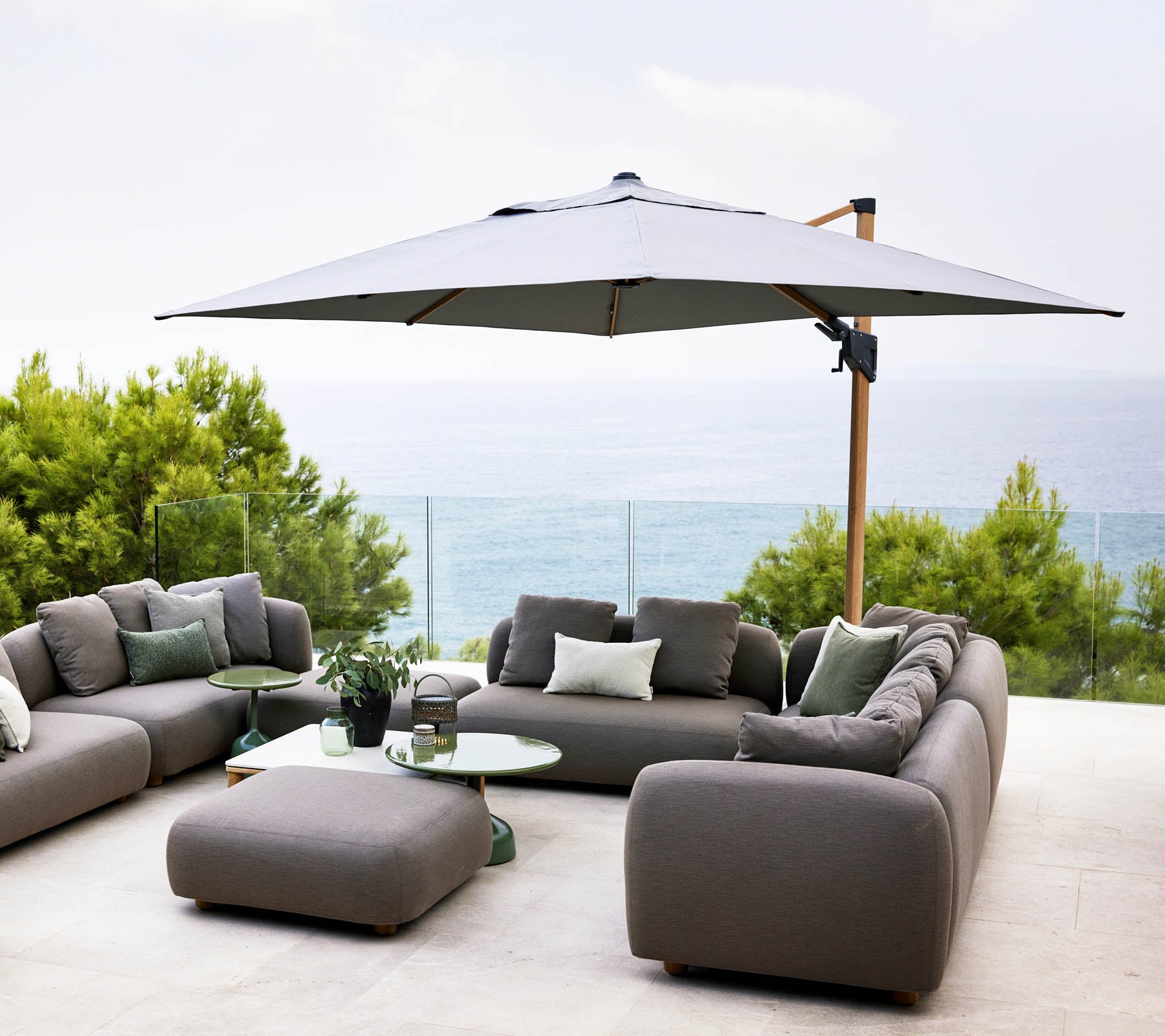 Stylischer Außenbereich mit großen Sonnenschirm, grauen Sofas und einem Glastisch mit Blick auf eine friedliche Wasseransicht und üppiges Grün.