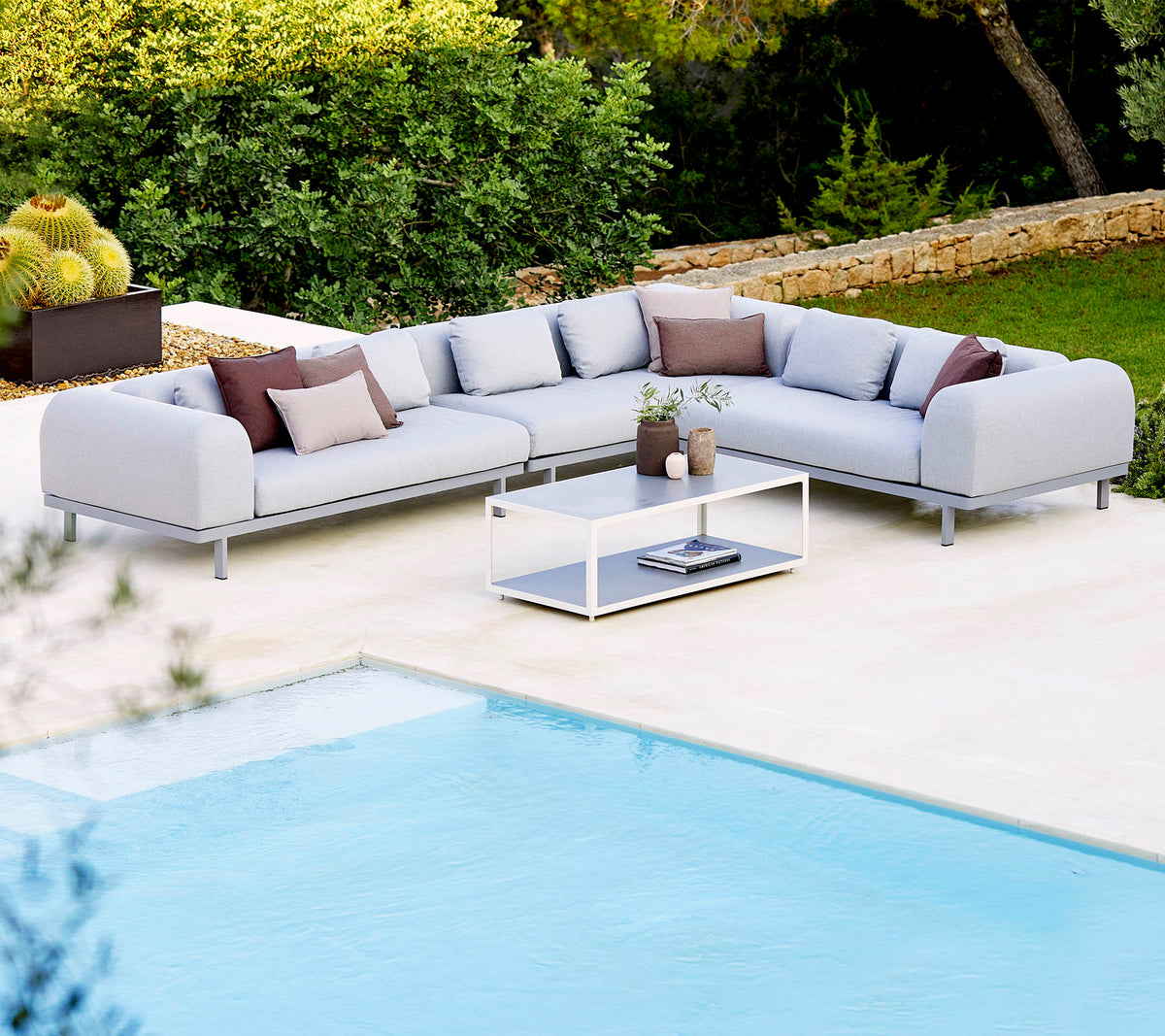Hellgraues Sectionalsofa mit dekorativen Kissen und einem Glastisch, der neben einem Swimmingpool und üppigem Grün positioniert ist.