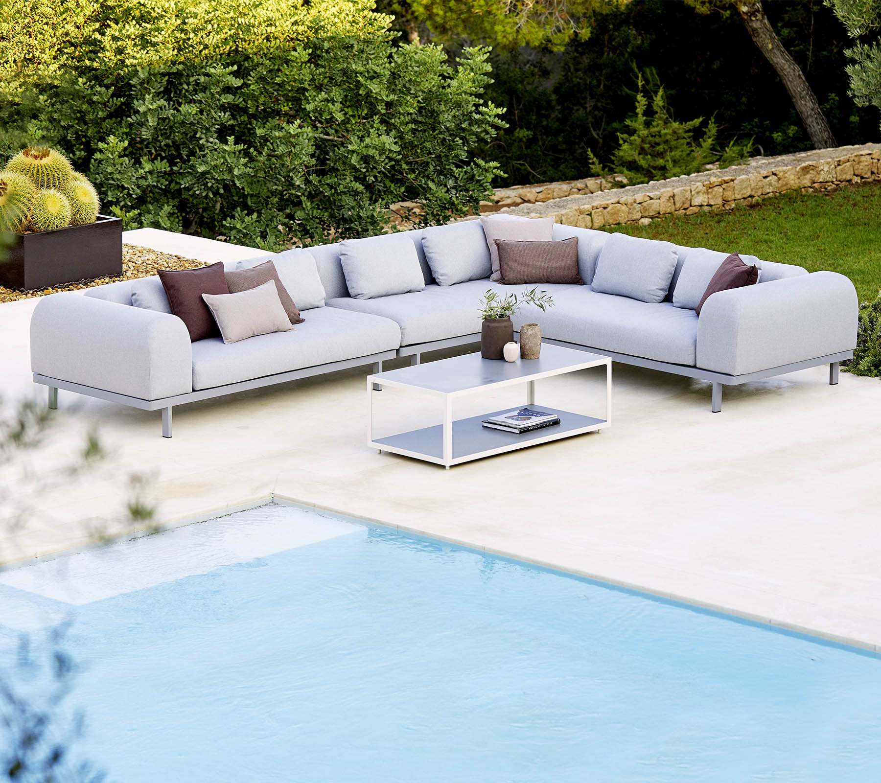 Hellgraues Sectionalsofa mit dekorativen Kissen und einem Glastisch, der neben einem Swimmingpool und üppigem Grün positioniert ist.
