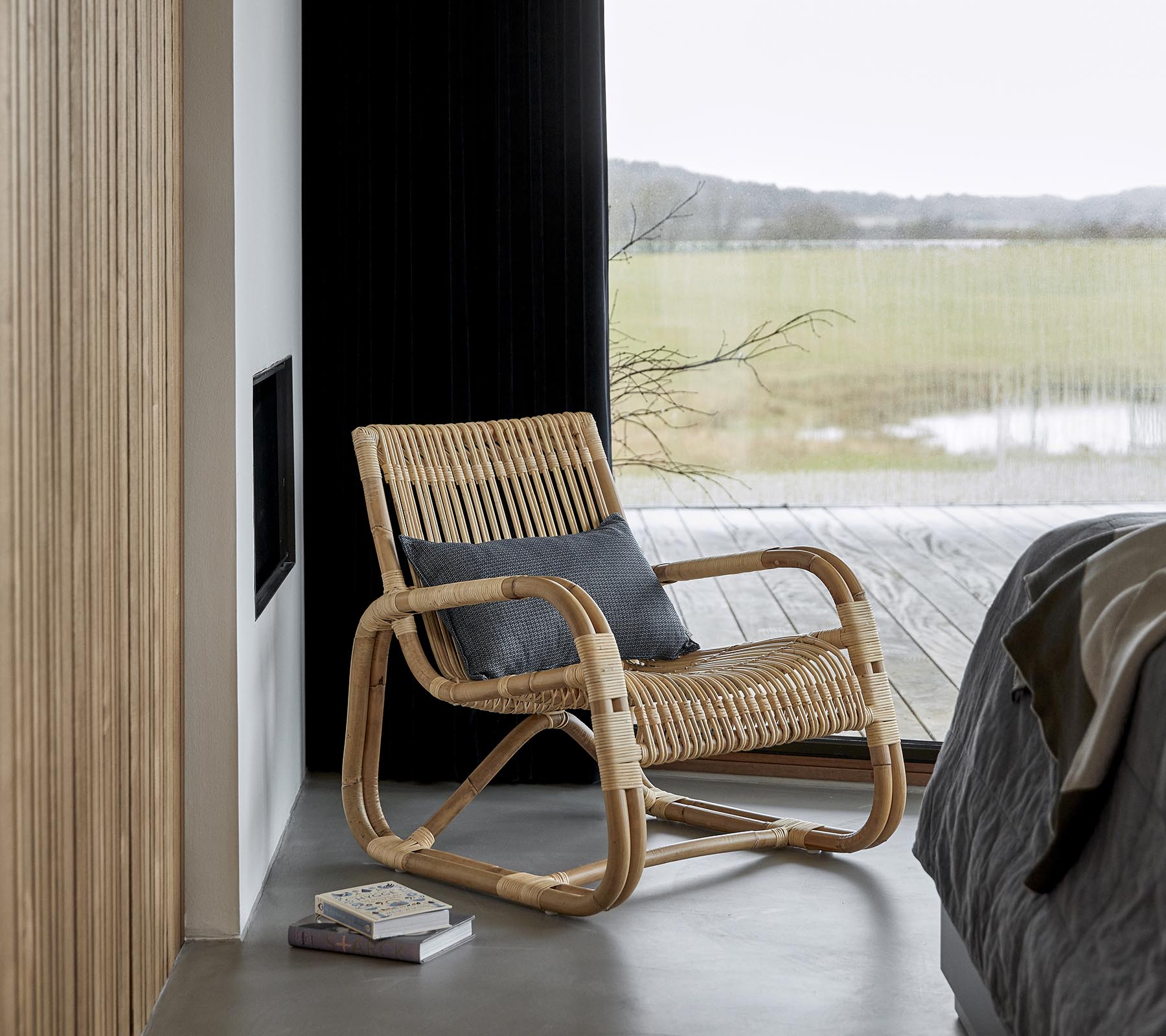 Ein stilvoller Schaukelstuhl aus natürlichen Materialien, neben einem Bett und einem Buch platziert, mit einem malerischen Blick, der durch große Fenster sichtbar ist.