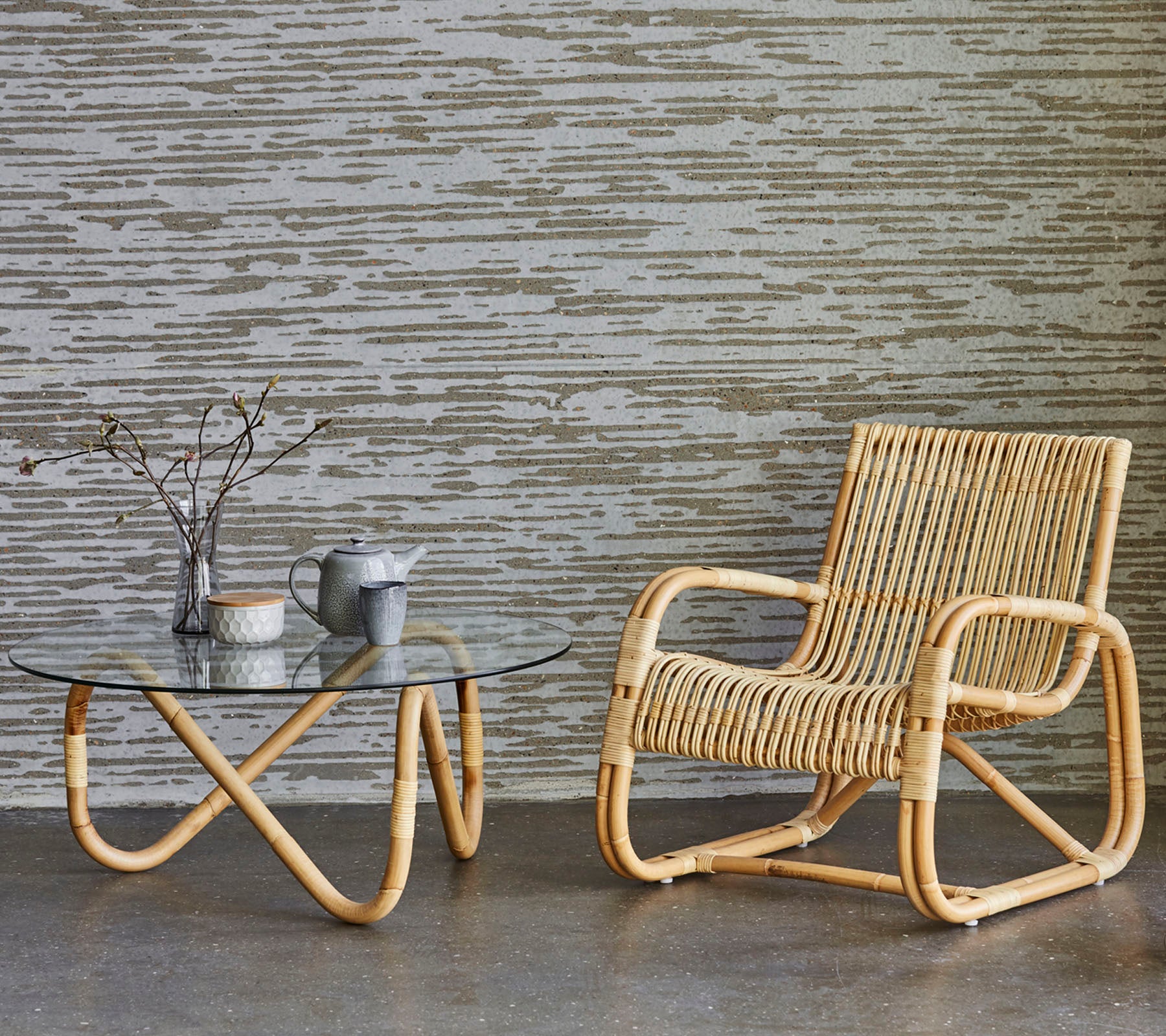 Rattansessel und Glastisch mit dekorativen Gegenständen, vor einer strukturierten Wand platziert. Ein minimalistischer und moderner Einrichtungsstil.