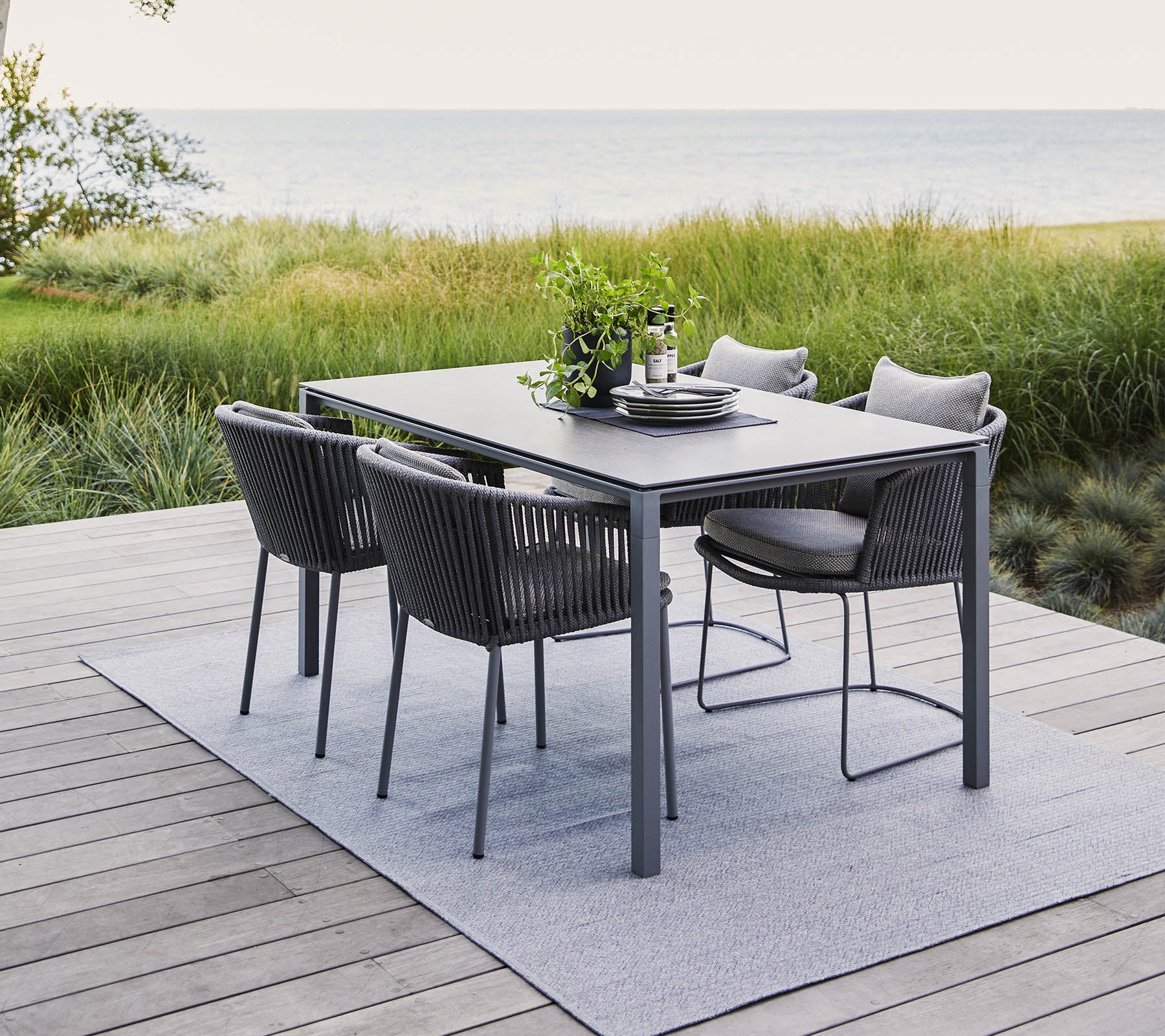 Ein modernes Outdoor-Essgruppen-Set mit einem rechteckigen Tisch und vier stilvollen Stühlen, umgeben von grünem Pflanzen und einem ruhigen Wasserblick.