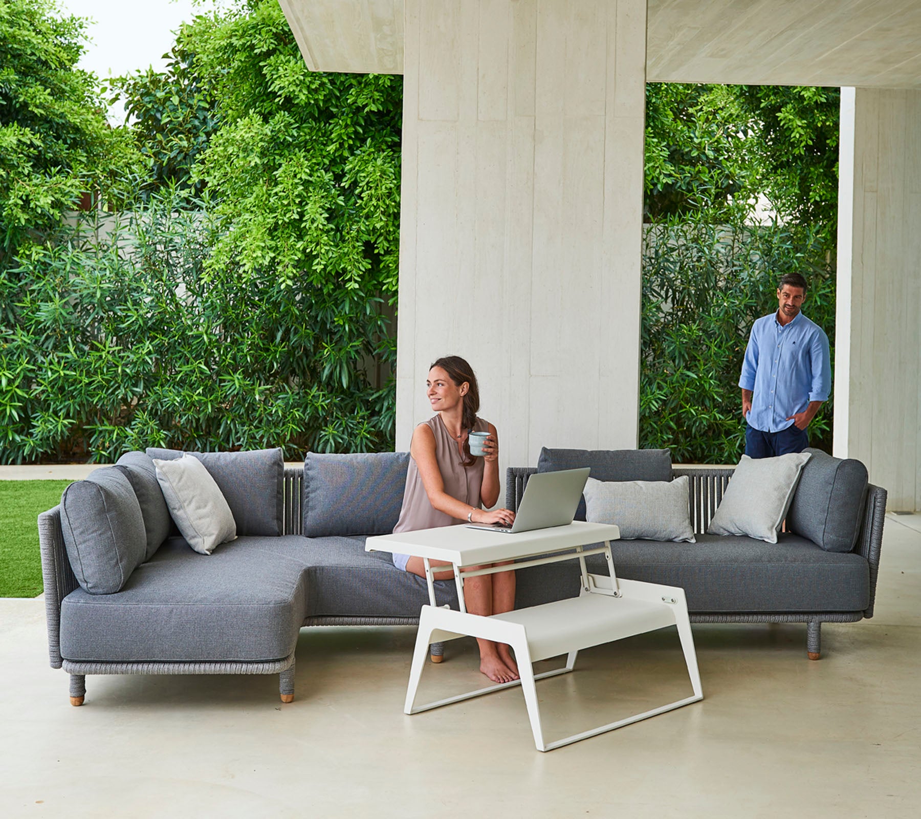 Ein stilvolles graues Ecksofa mit Kissen, begleitet von einem modernen Couchtisch, und einer Frau, die an einem Laptop arbeitet, während ein Mann in der Nähe steht.