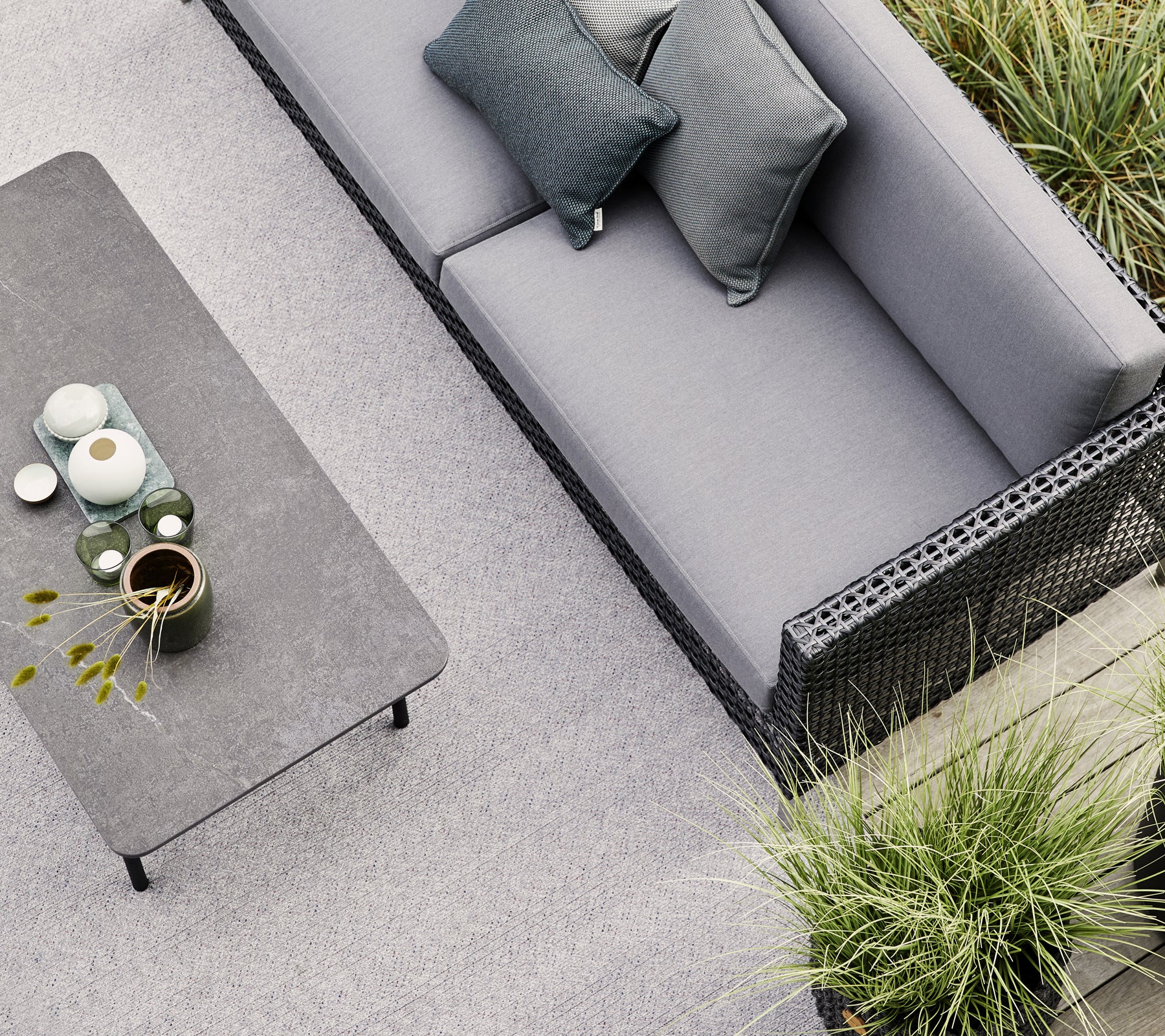 Ein graues Sofa mit zwei Kissen steht neben einem minimalistischen Tisch, der mit Keramiken und einer kleinen Pflanze geschmückt ist, umgeben von Grün.