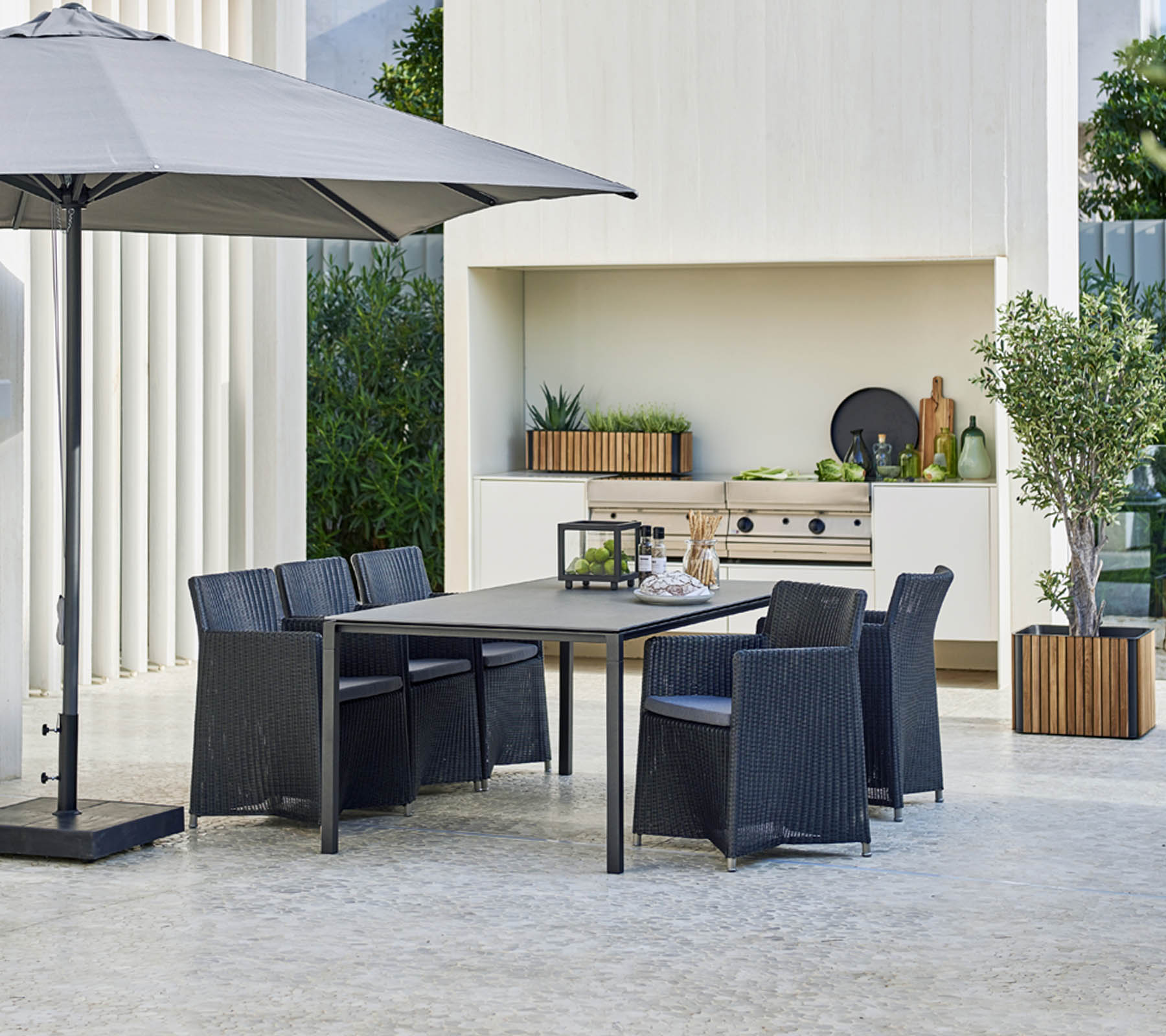 Modernes Outdoor-Esstischset mit einem großen Tisch, sechs Stühlen, einem stilvollen Sonnenschirm und einem Hintergrund mit Grün und dekorativen Elementen.