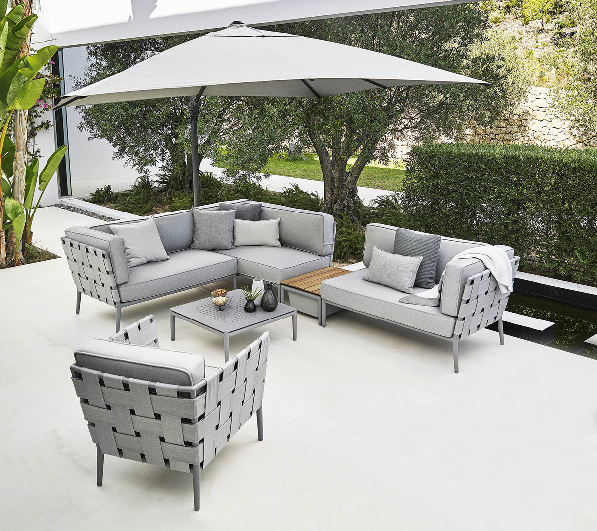 Stylische Outdoor-Sitzgelegenheit mit einem grauen Ecksofa, einem Sessel und einem Couchtisch unter einem großen Sonnenschirm.