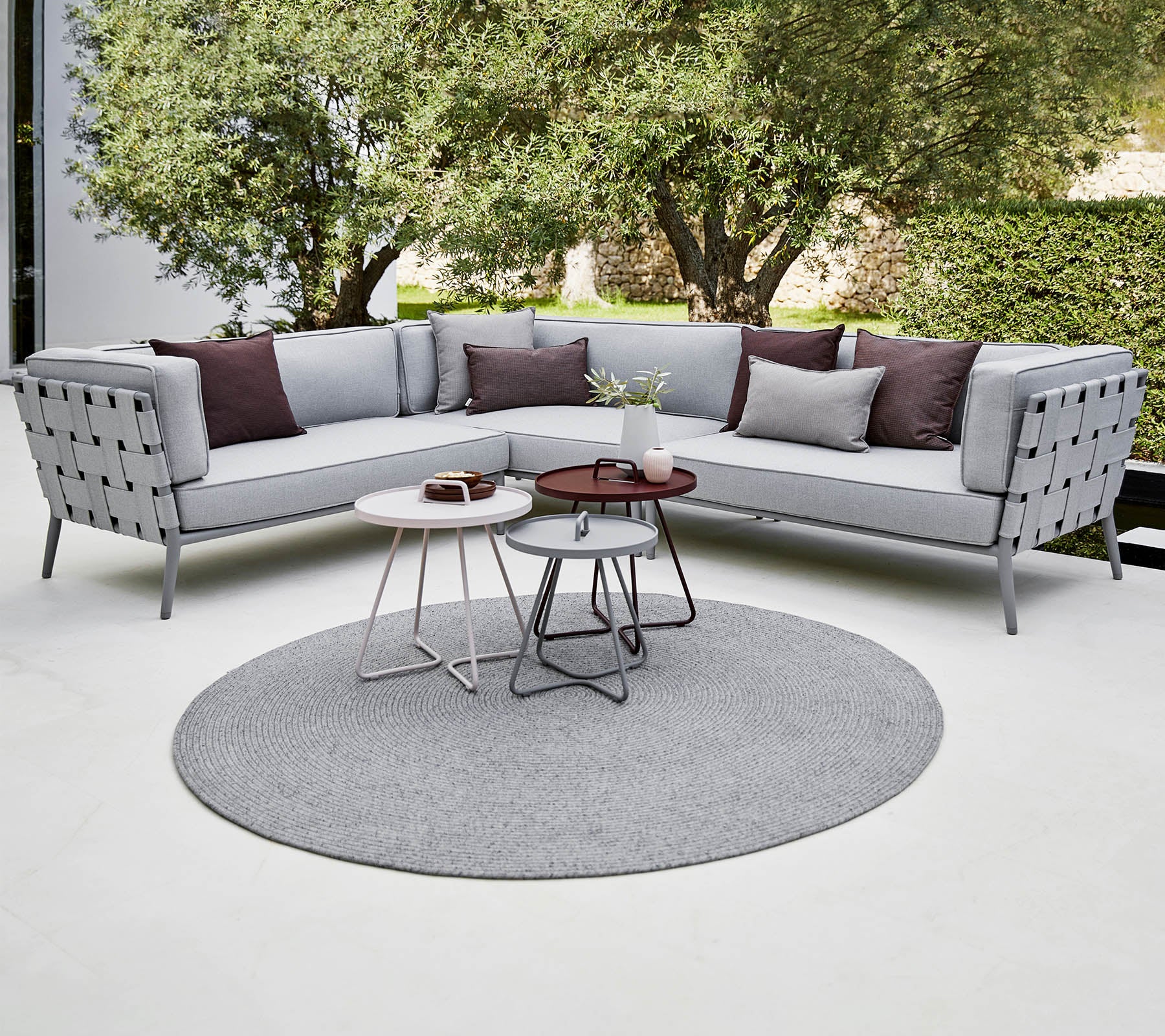 Moderne Ecksofa in Hellgrau mit dekorativen Kissen, begleitet von stilvollen Couchtischen auf einem runden Teppich. Üppiges Grün im Hintergrund.