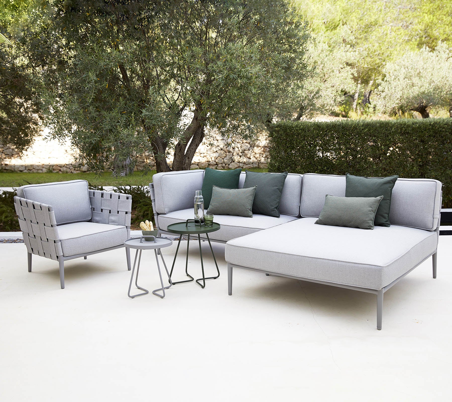 Moderne Außen-Sitzgelegenheit mit einer sectional Sofa mit Kissen, einem Stuhl und einem kleinen Glastisch, umgeben von Grün.