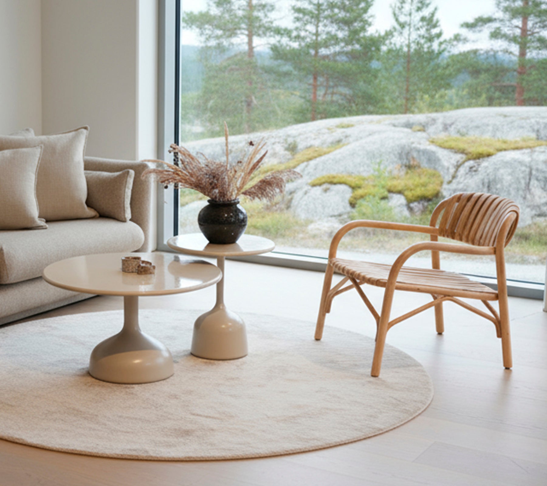 Moderne Lounge mit einem Sofa, einem runden Tisch, einer schwarzen Vase und einem Rattanstuhl neben einem großen Fenster.