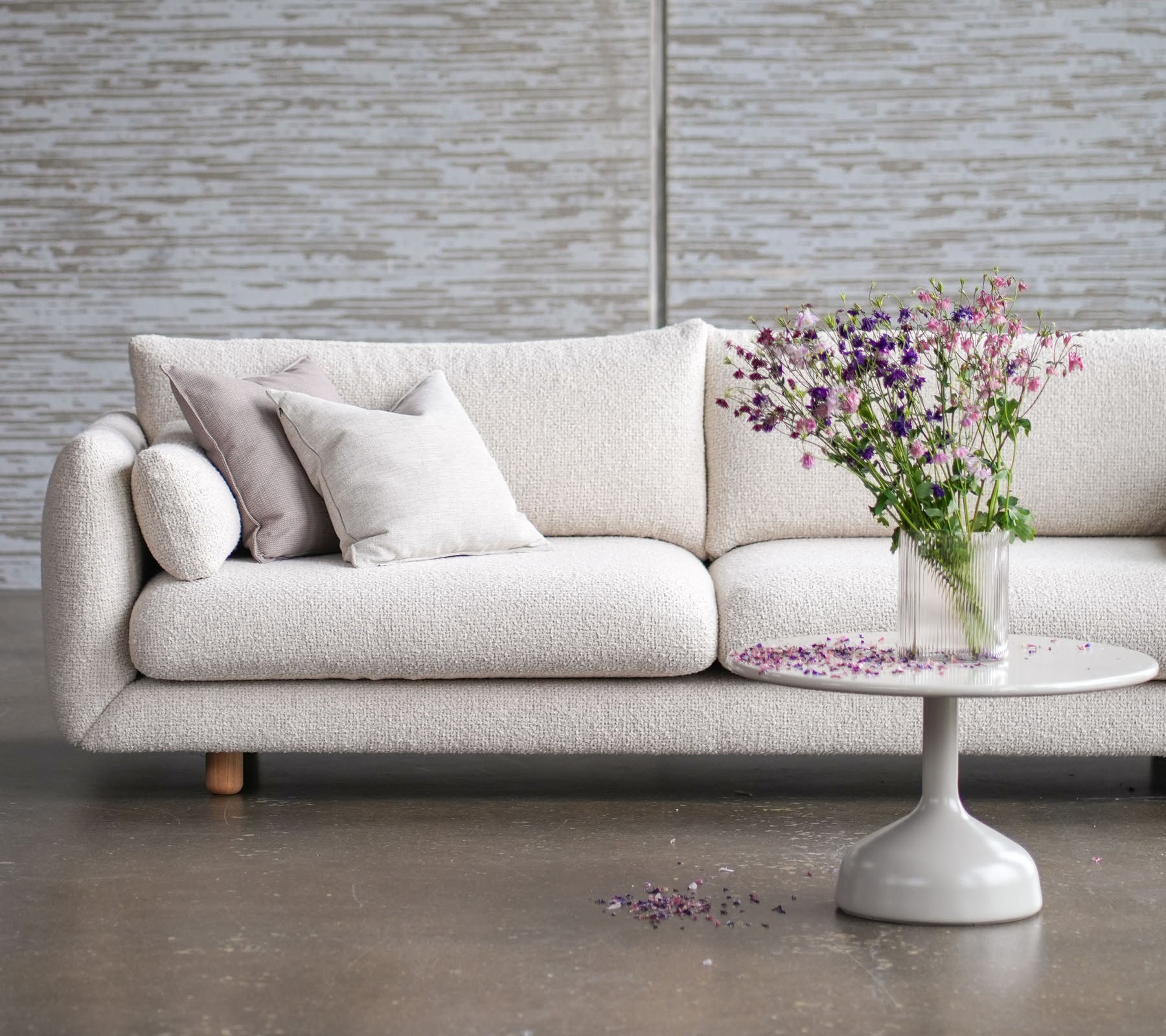 Helle Couch mit Teakholzbeinen, geschmückt mit Kissen, ergänzt durch einen runden Tisch und eine Vase mit frischen Blumen.