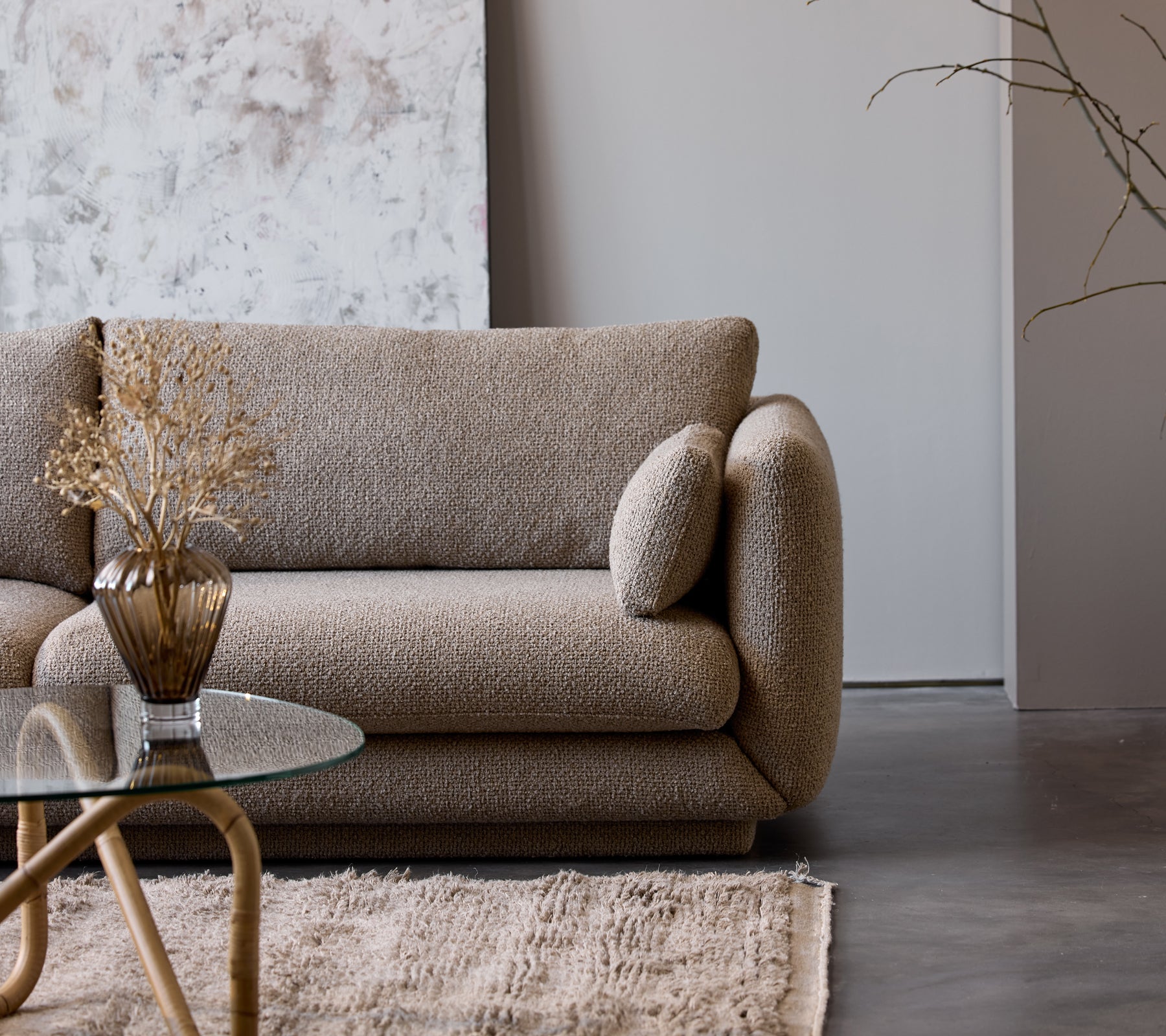 Ein taupefarbenen Couchtisch mit einer Glasplatte steht neben einem strukturierten beigen Sofa, geschmückt mit einer dekorativen Vase und getrockneten Blumen.