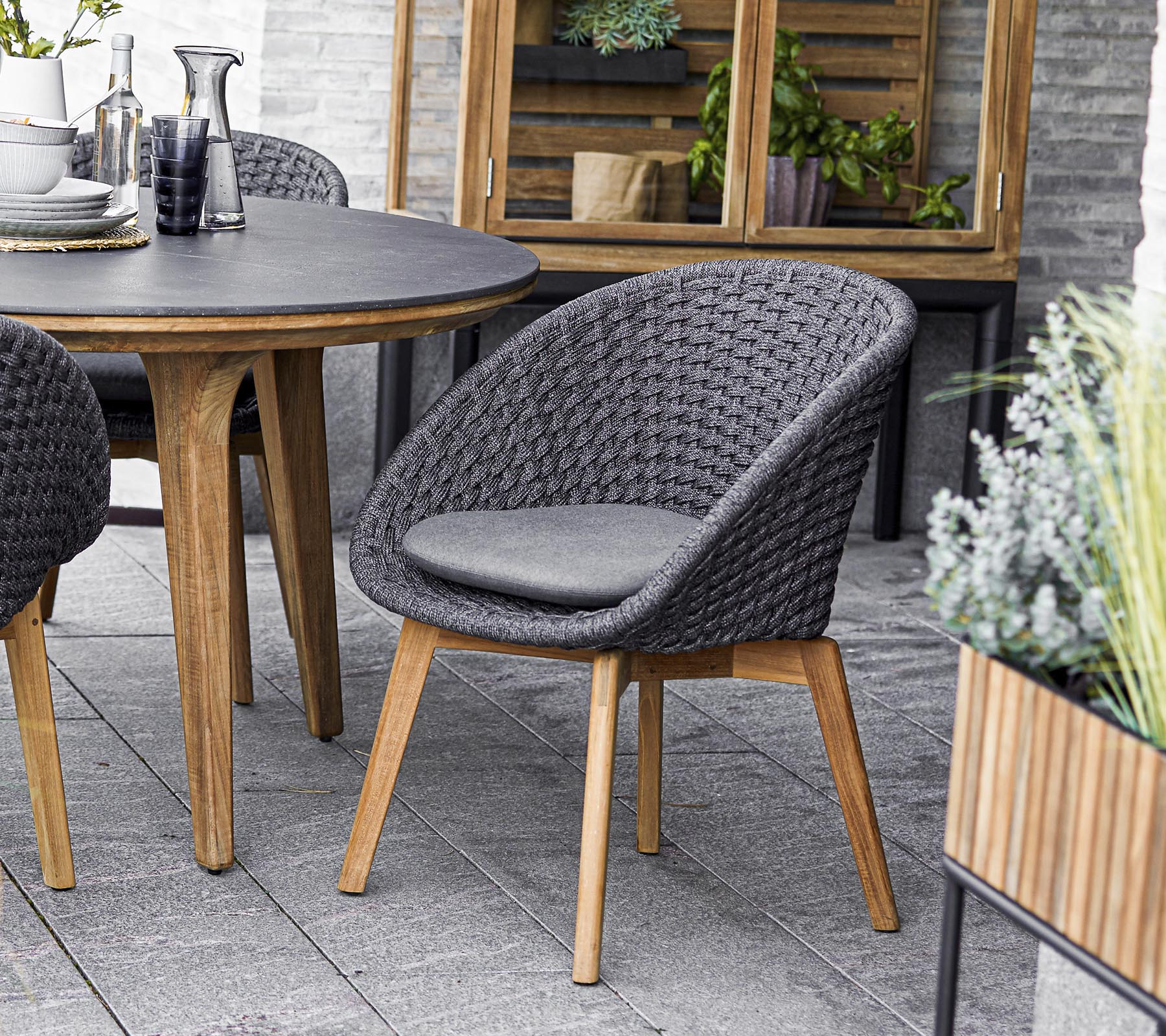 Stylischer geflochtener Stuhl mit gepolsterter Sitzfläche, kombiniert mit einem runden Tisch und umgeben von Grünpflanzen. Perfekt für gesellige Zusammenkünfte.
