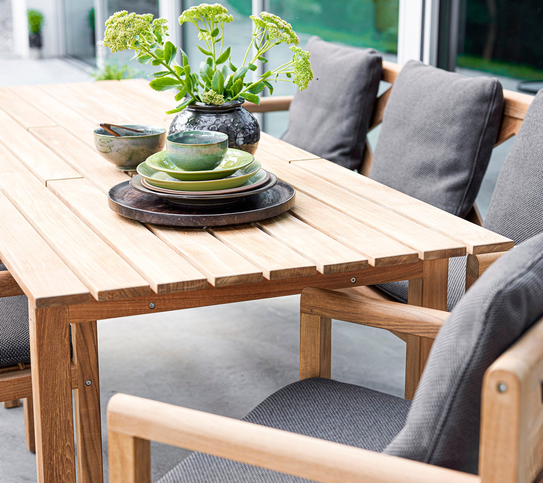 Stilvolles Holztisch-Set mit Kissen, Tellern und Schalen, akzentuiert durch eine Vase mit Grünpflanzen. Perfekt für einladende Zusammenkünfte.