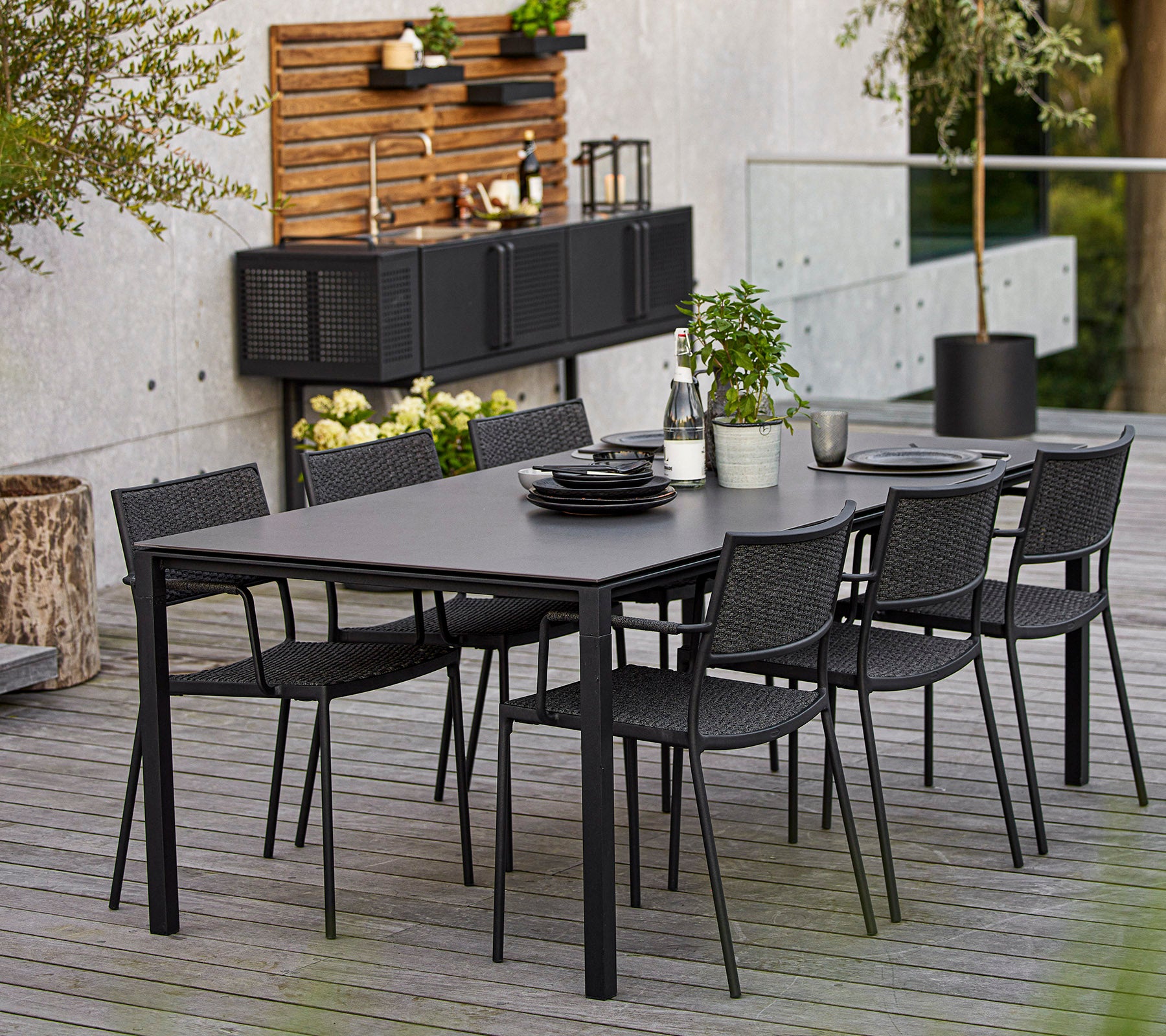 Ein modernes Esszimmer-Set mit einem großen rechteckigen Tisch und sechs schwarzen geflochtenen Stühlen, akzentuiert durch dekorative Elemente und Grünpflanzen.