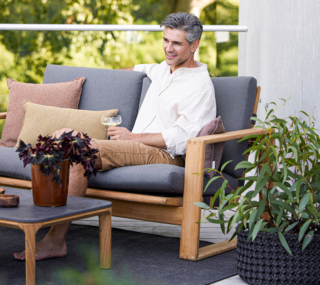 Ein entspannter Mann sitzt auf einem stilvollen Sofa mit Kissen und genießt ein Getränk neben einer Topfpflanze und einem Couchtisch.