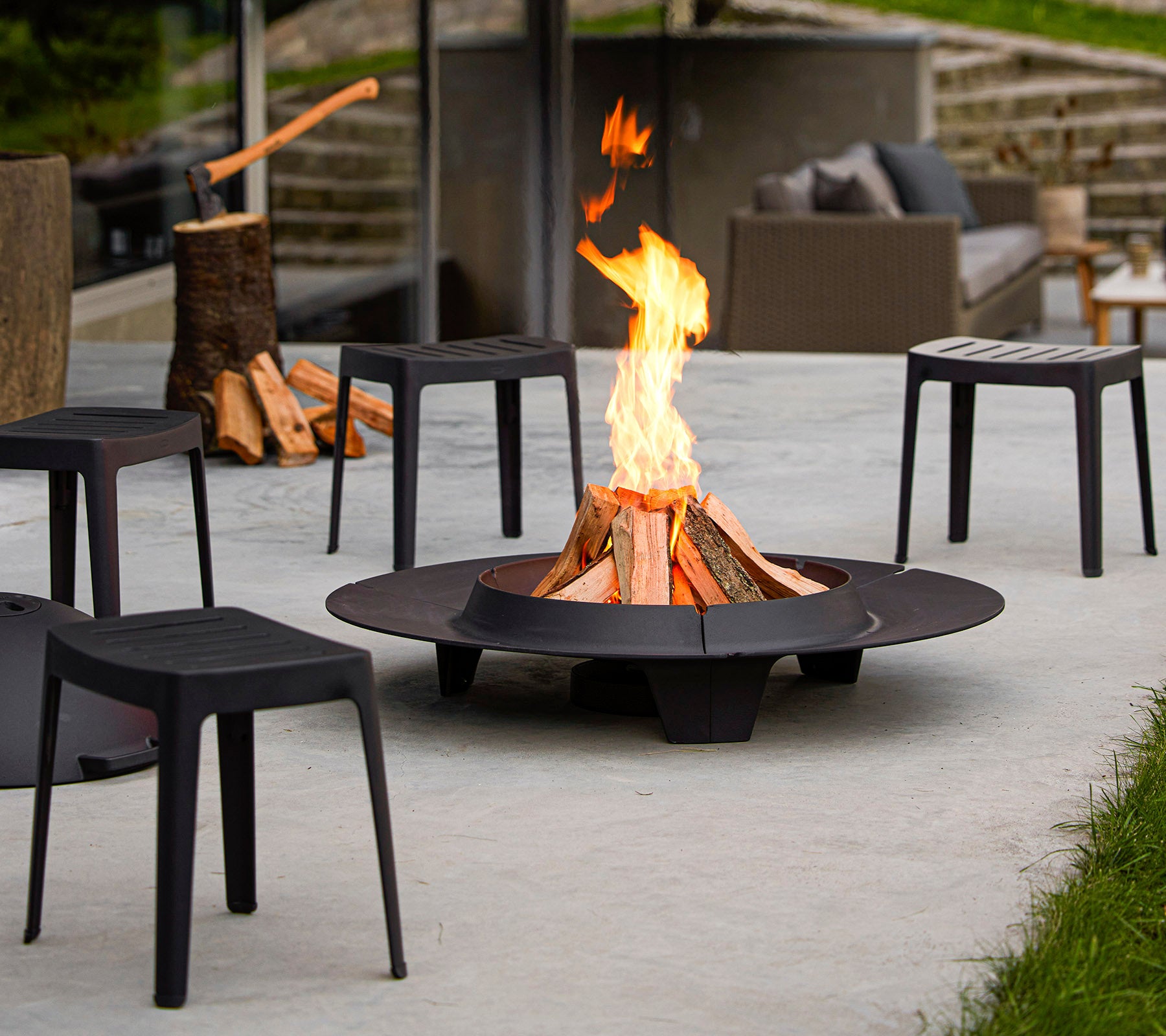 Ein Feuerstelle umgeben von minimalistischen schwarzen Stühlen, mit einer lebhaften Flamme und Holzstapeln in der Nähe.