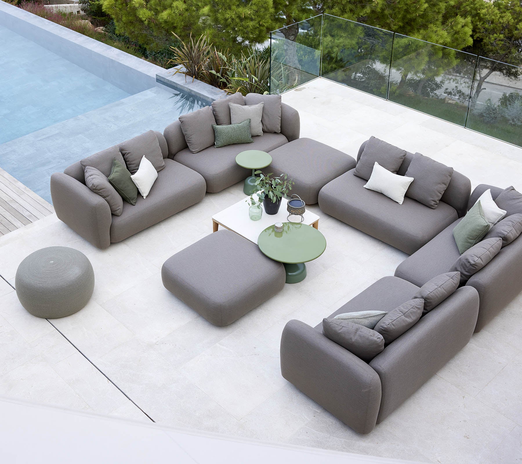 Moderne Outdoor-Sitzarrangement mit grauen Sofas und grünen Tischen neben einem Pool, umgeben von Grün.