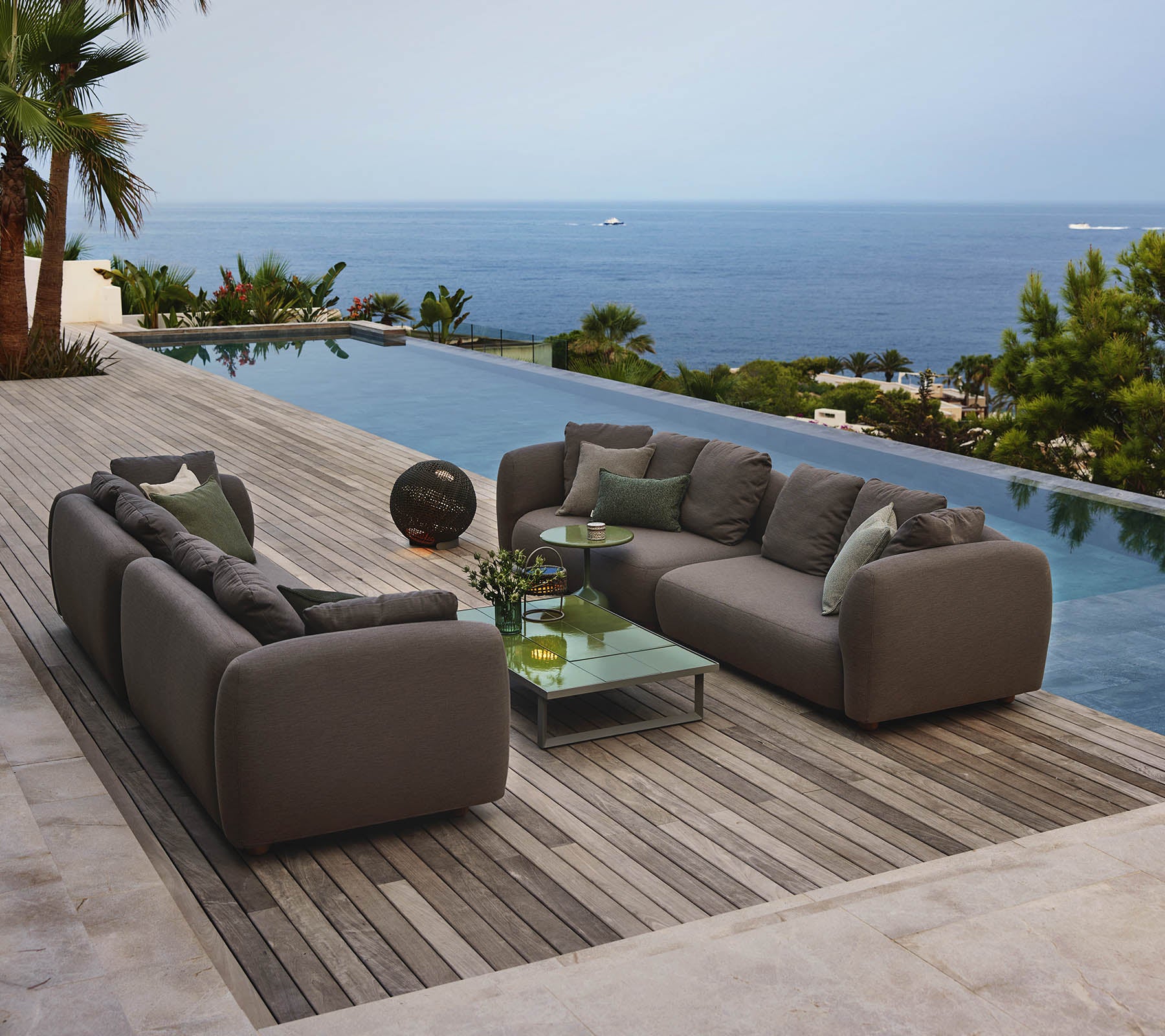Moderne Loungemöbel, die um einen Glastisch angeordnet sind, mit Blick auf einen ruhigen Pool und einen weiten Ozeanblick, umgeben von Grün.